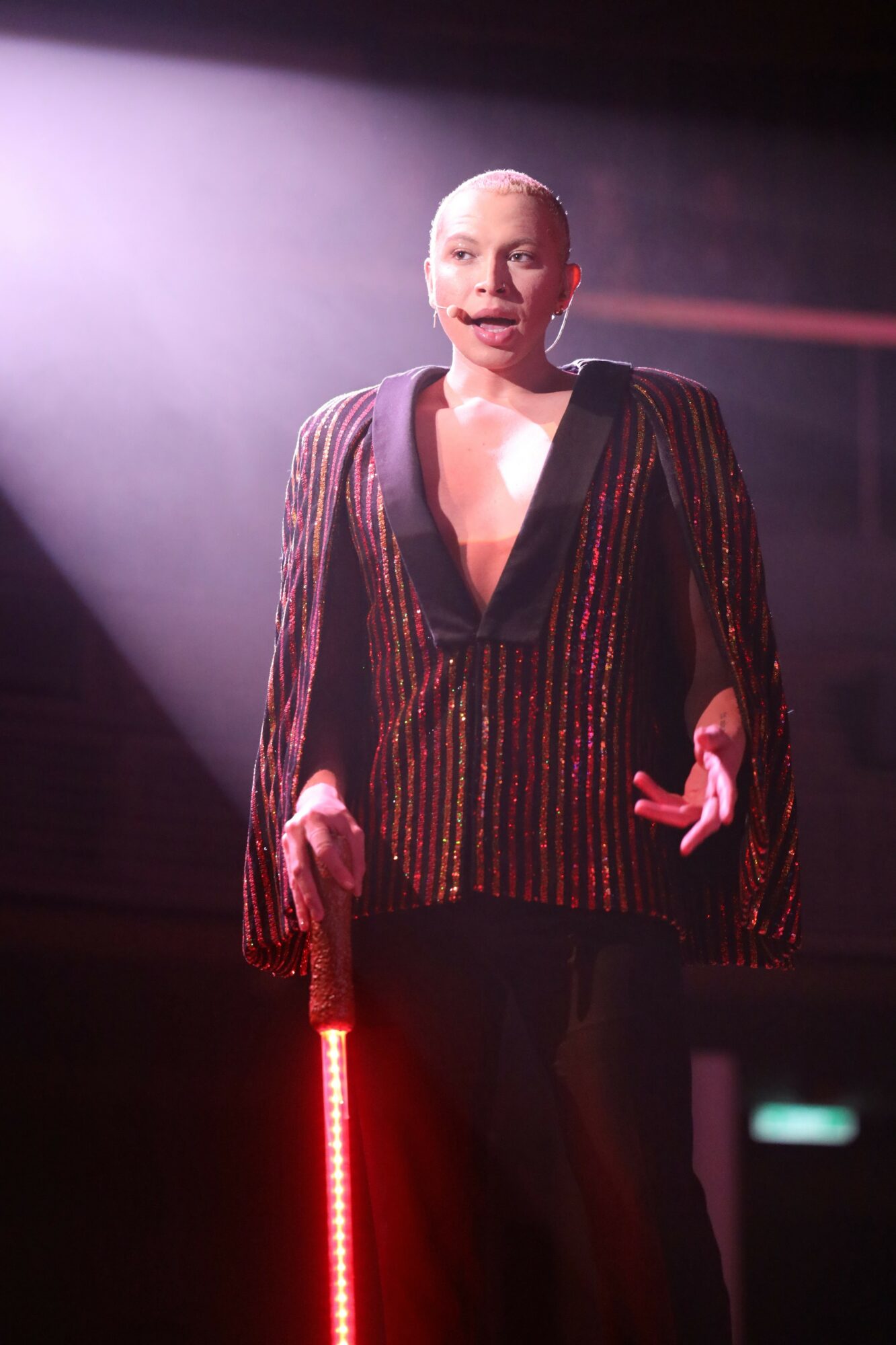Person standing on stage with spotlight, wearing a striped blazer, holding a red light stick, speaking or performing.