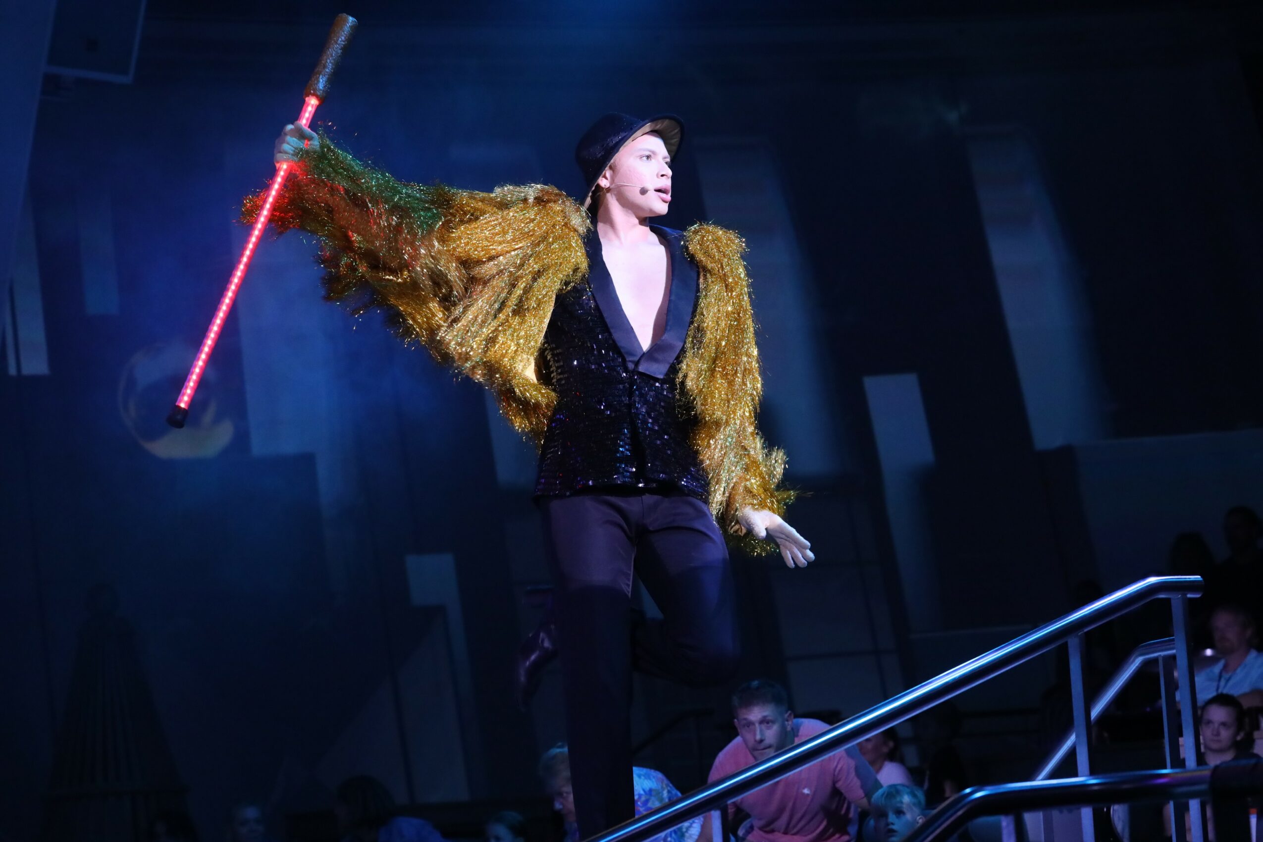Performer on stage holding a red and white baton, wearing a gold jacket and black outfit, with a dark background.