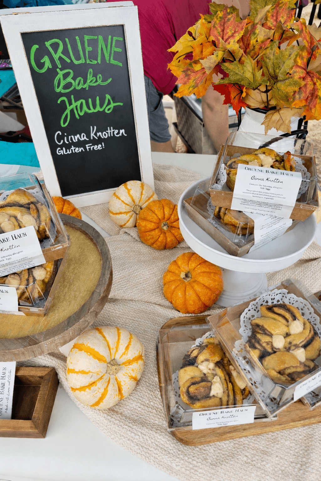 Display of baked goods with small pumpkins, autumn leaves, and a chalkboard sign reading 'Gluene Bae Haus' and 'Cinna Knotten Gluten Free'.