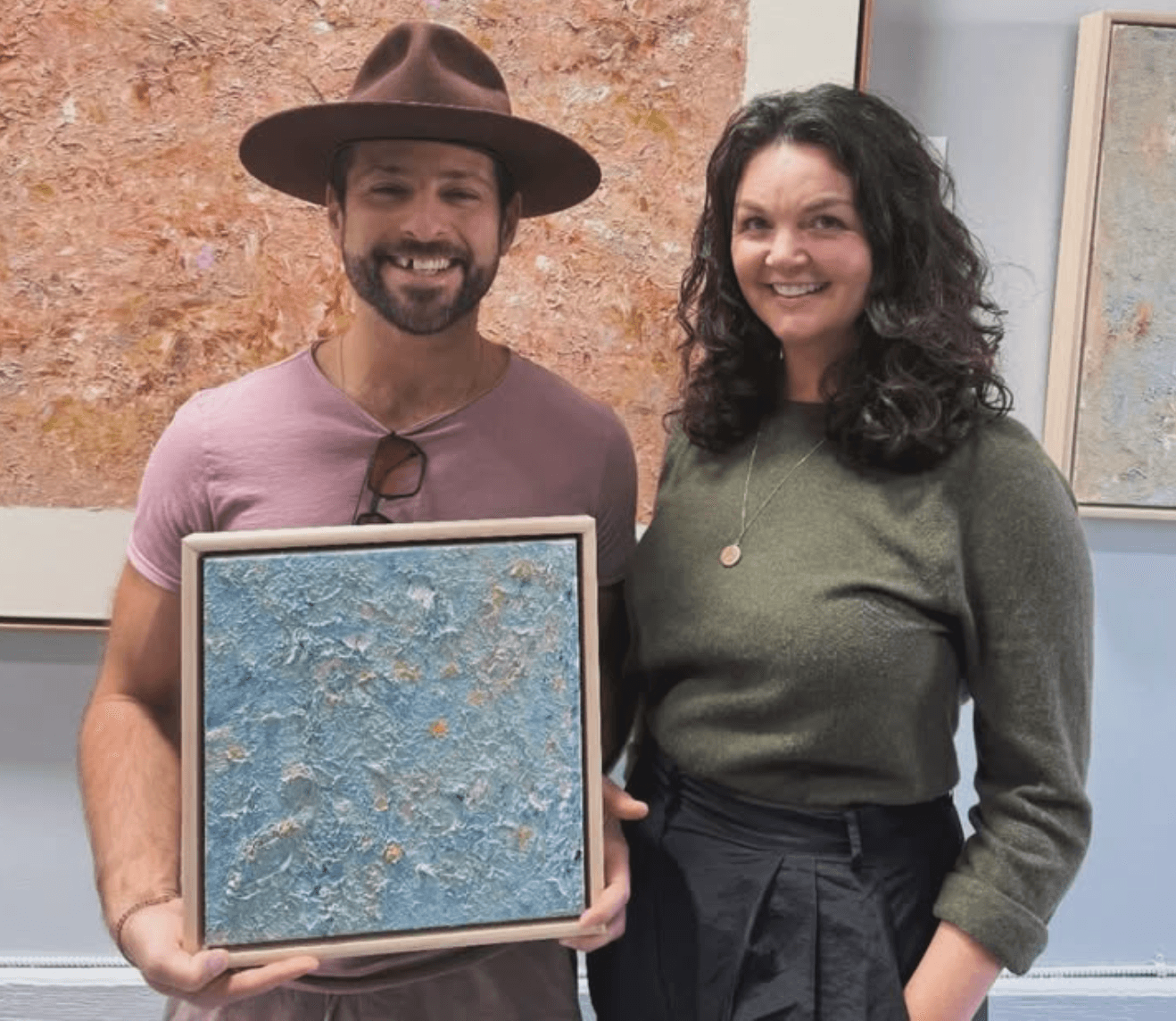 Two people standing indoors, one holding a framed textured artwork, both smiling, with a textured wall behind them.