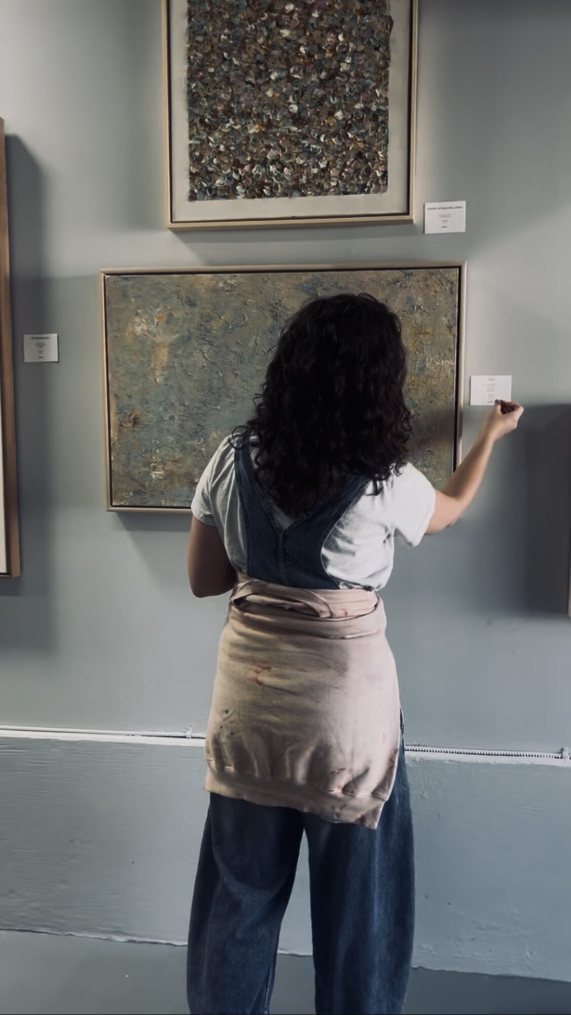 Person with curly hair wearing a white shirt and beige skirt, facing artwork on wall, holding a phone.