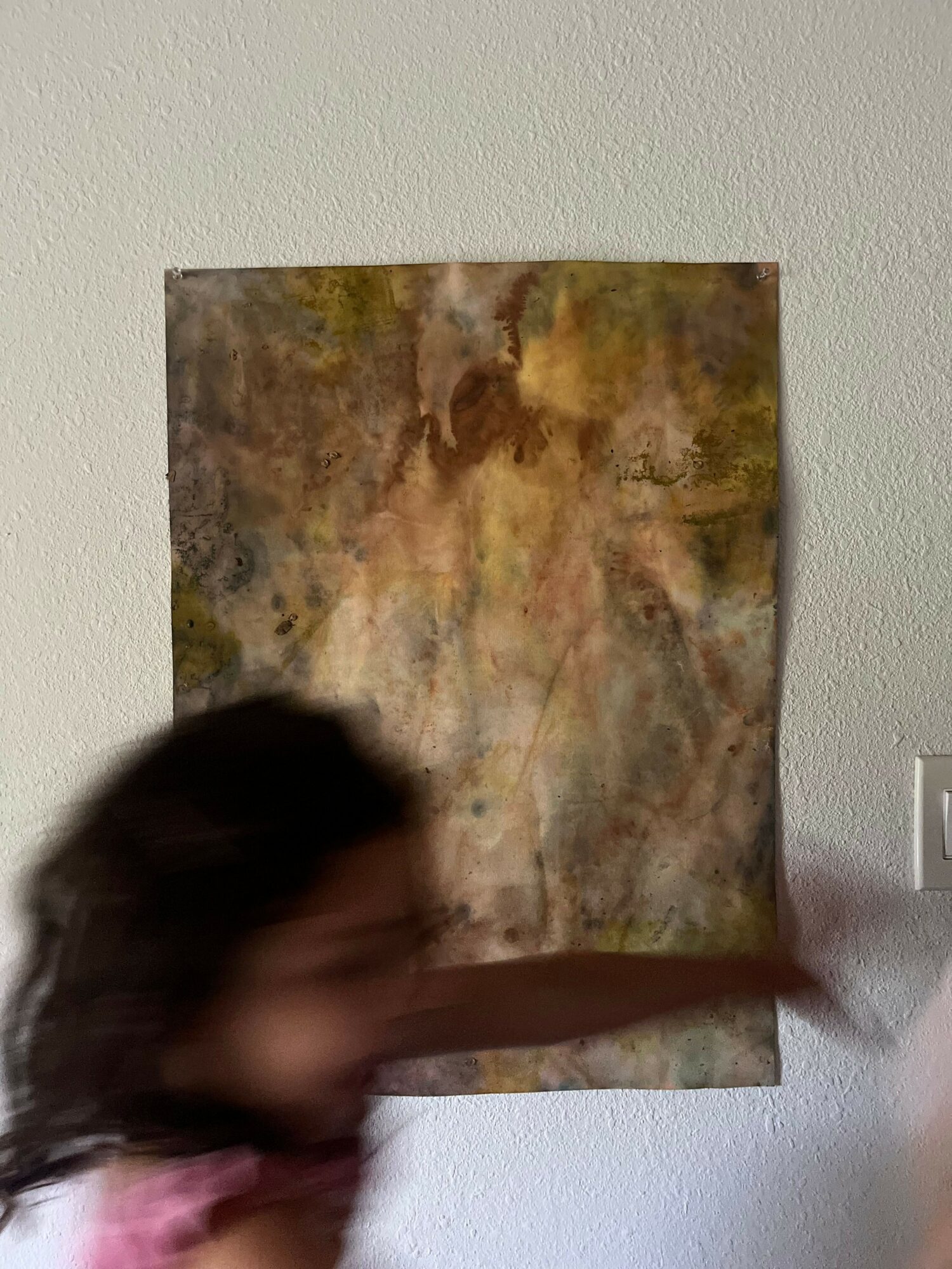 Blurry person with dark hair and pink shirt in front of a textured, multicolored wall.