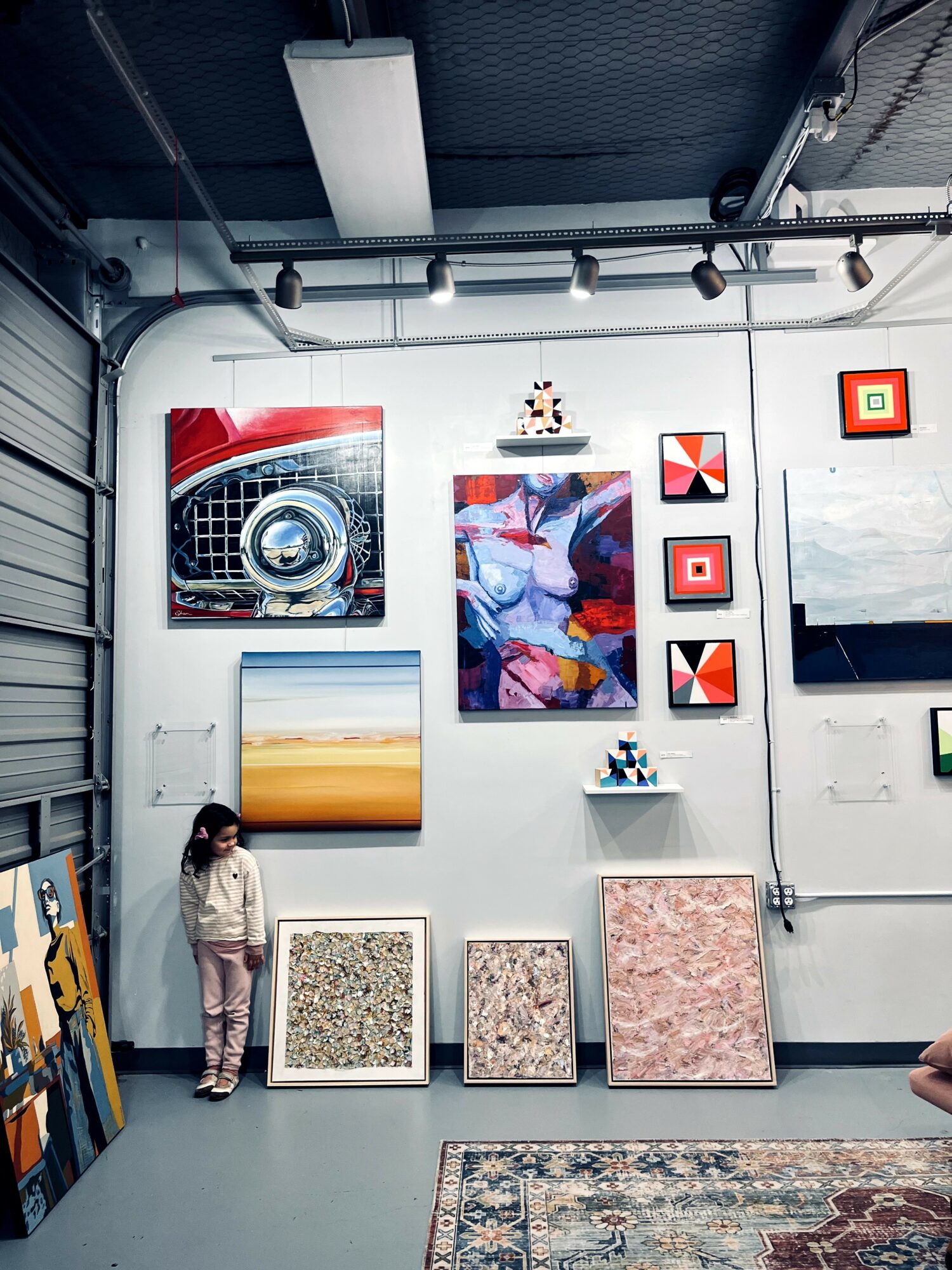 Child standing in an art gallery with various colorful paintings and artworks on the wall behind.