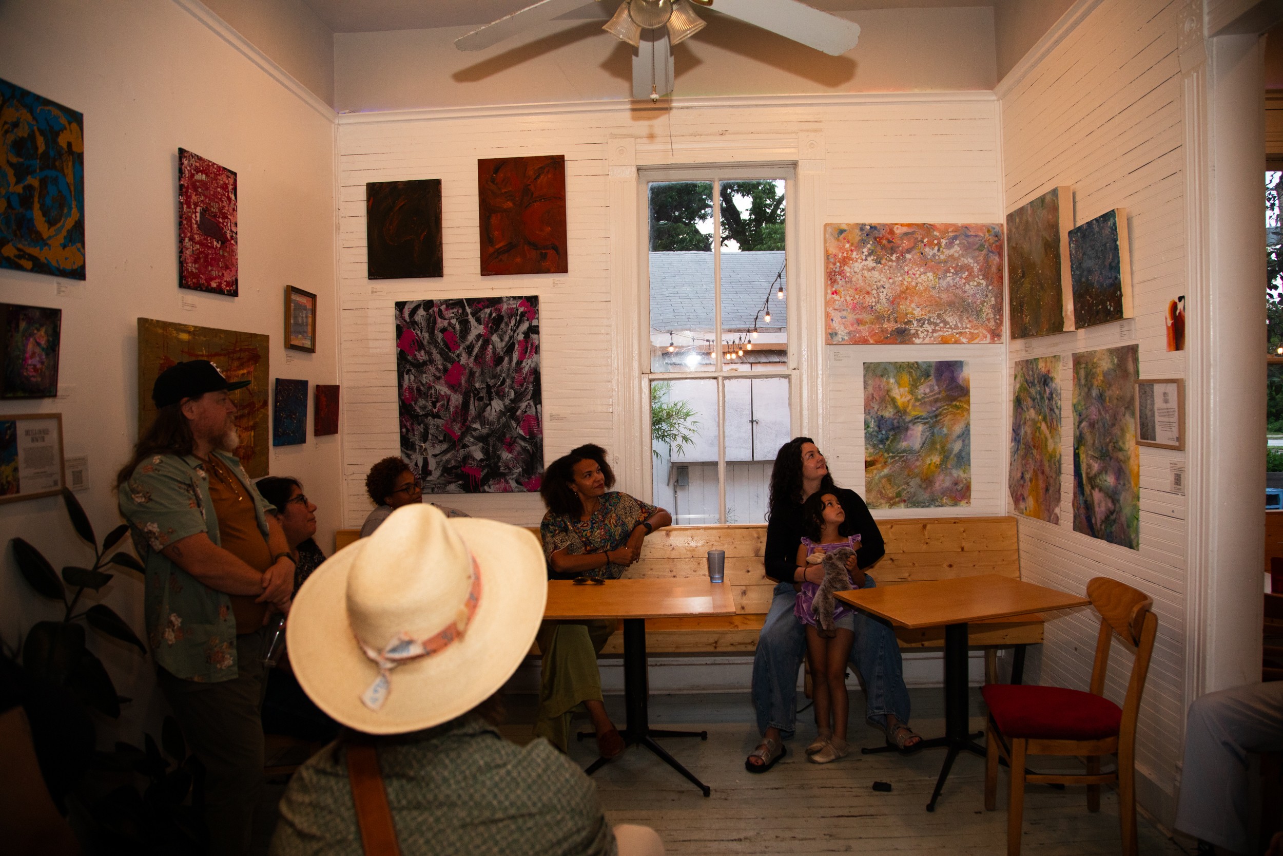 People viewing artwork in a gallery with paintings on white walls and a window in the background.