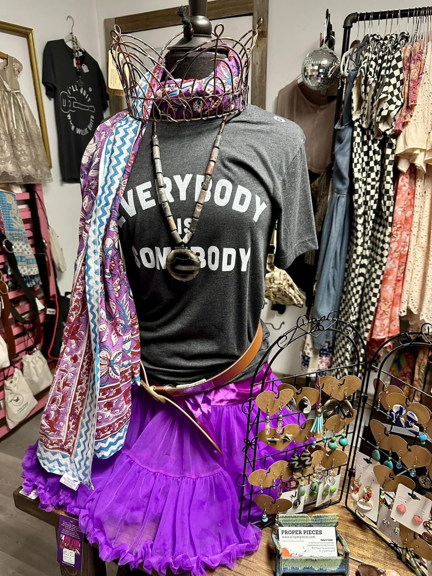 Mannequin wearing a black T-shirt with white text, purple tutu, and accessories, surrounded by clothing and accessories in a boutique.