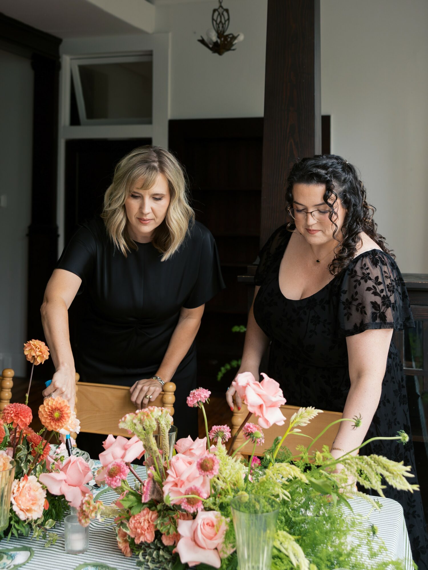 Two women stand behind a table with pink and orange flowers, indoors with a dark wooden beam and window in background.