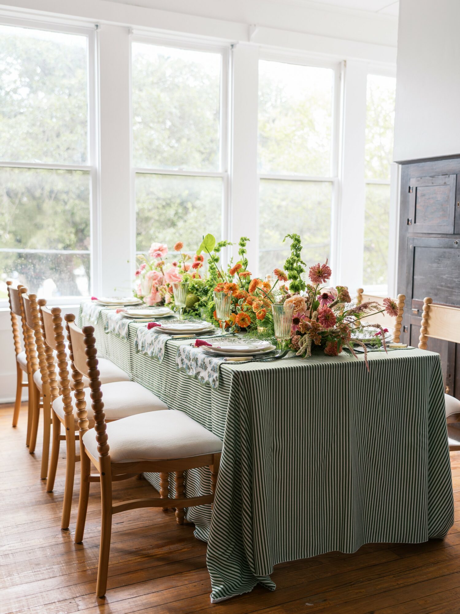 Long dining table with floral arrangements, set with plates and silverware, in a bright room with large windows.