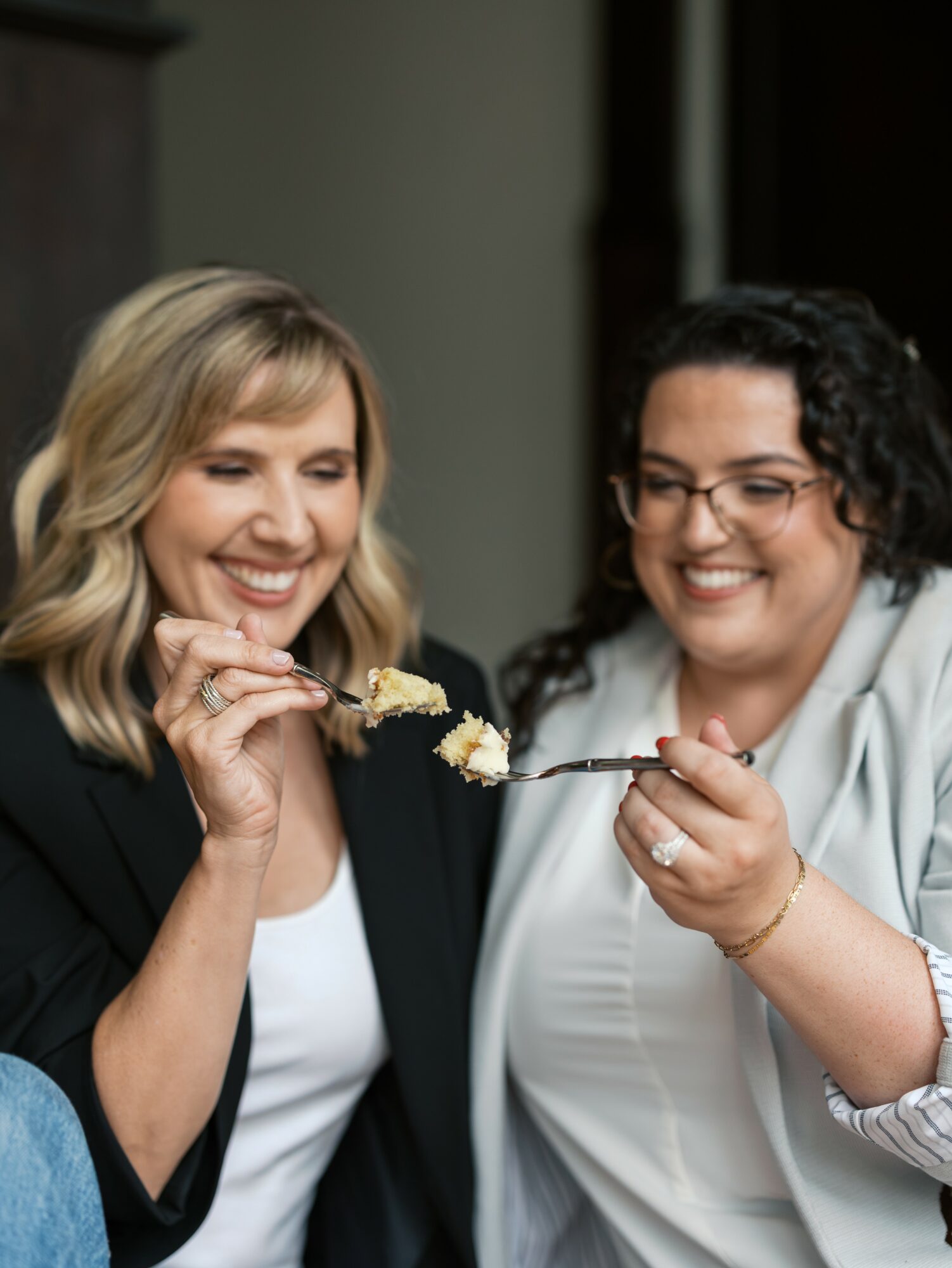 Two women smiling and sharing a fork with cake indoors.