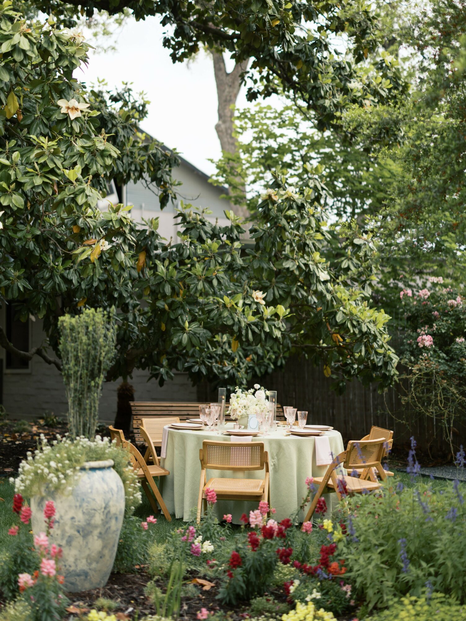 Outdoor garden with a round table, chairs, and lush greenery, including trees and flowering plants.