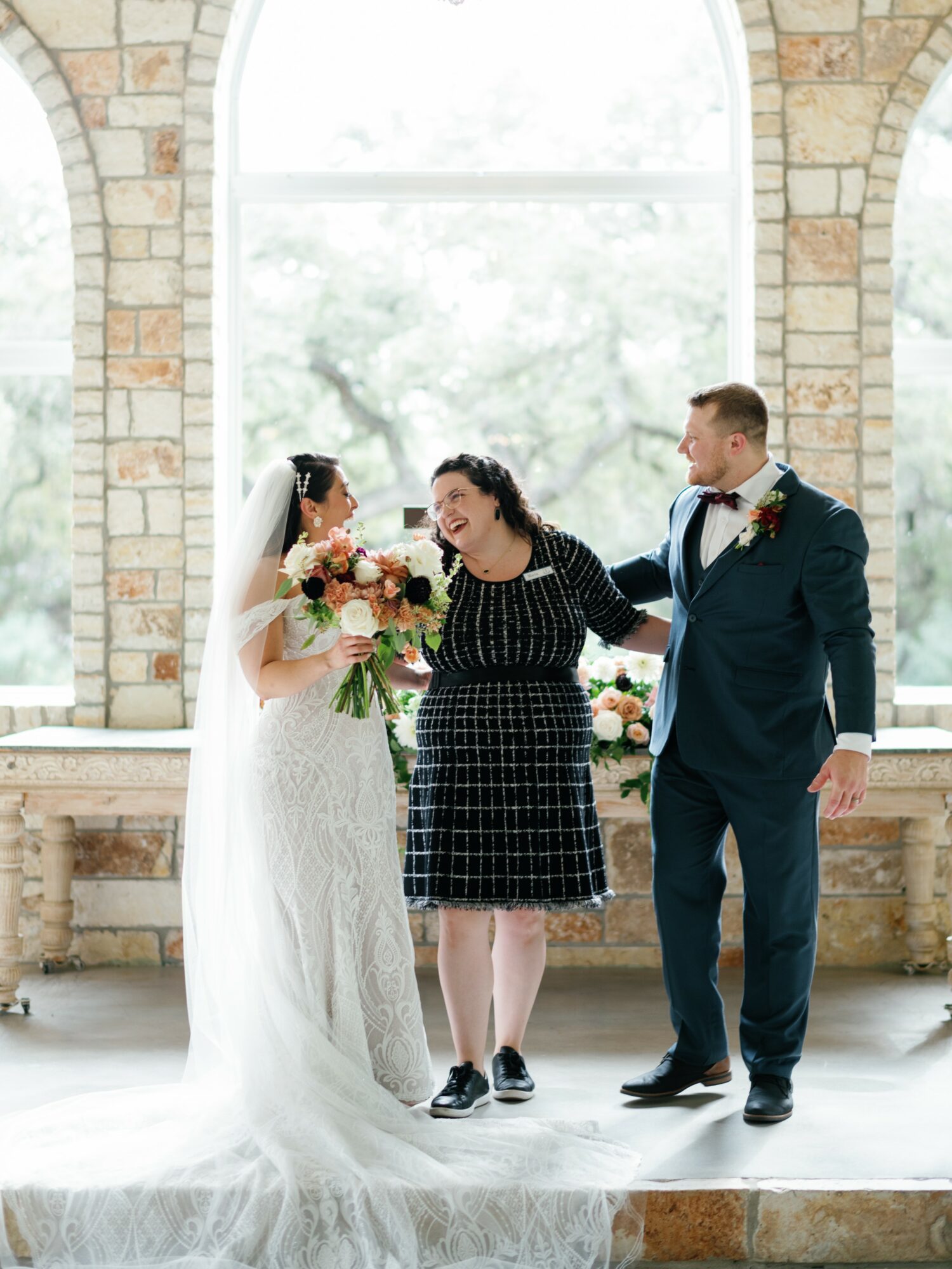 Bride, groom, and a woman stand together inside a bright room with large arched windows and brick walls, holding flowers.