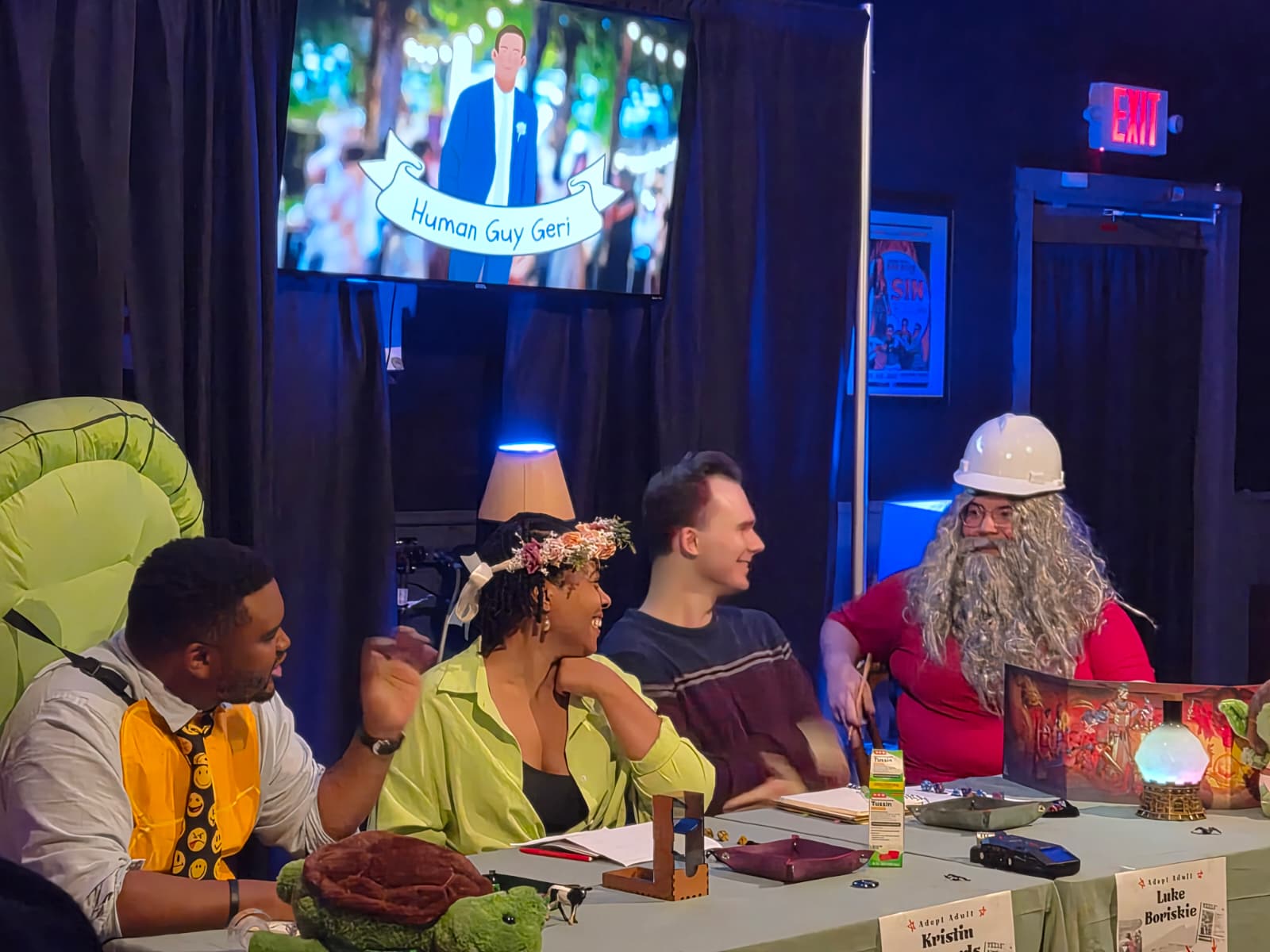 Four people sitting at a table with a person dressed as a wizard, a woman with a flower crown, and two men, in a room with a TV screen and decorations.