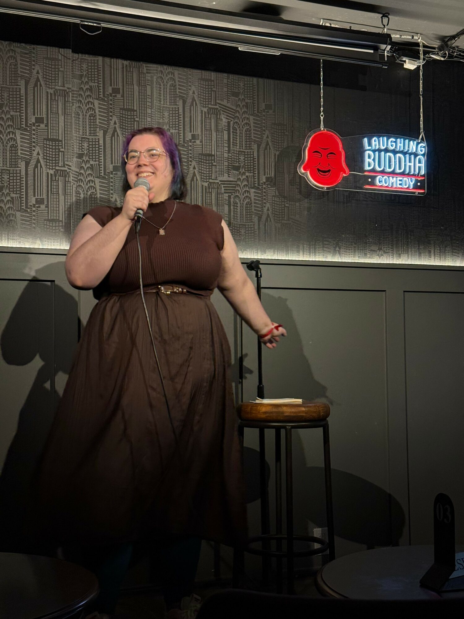 Person with purple hair taking a mirror selfie in a room with a sign that says 'BUDDING' and a red skull logo, holding a phone to their ear.