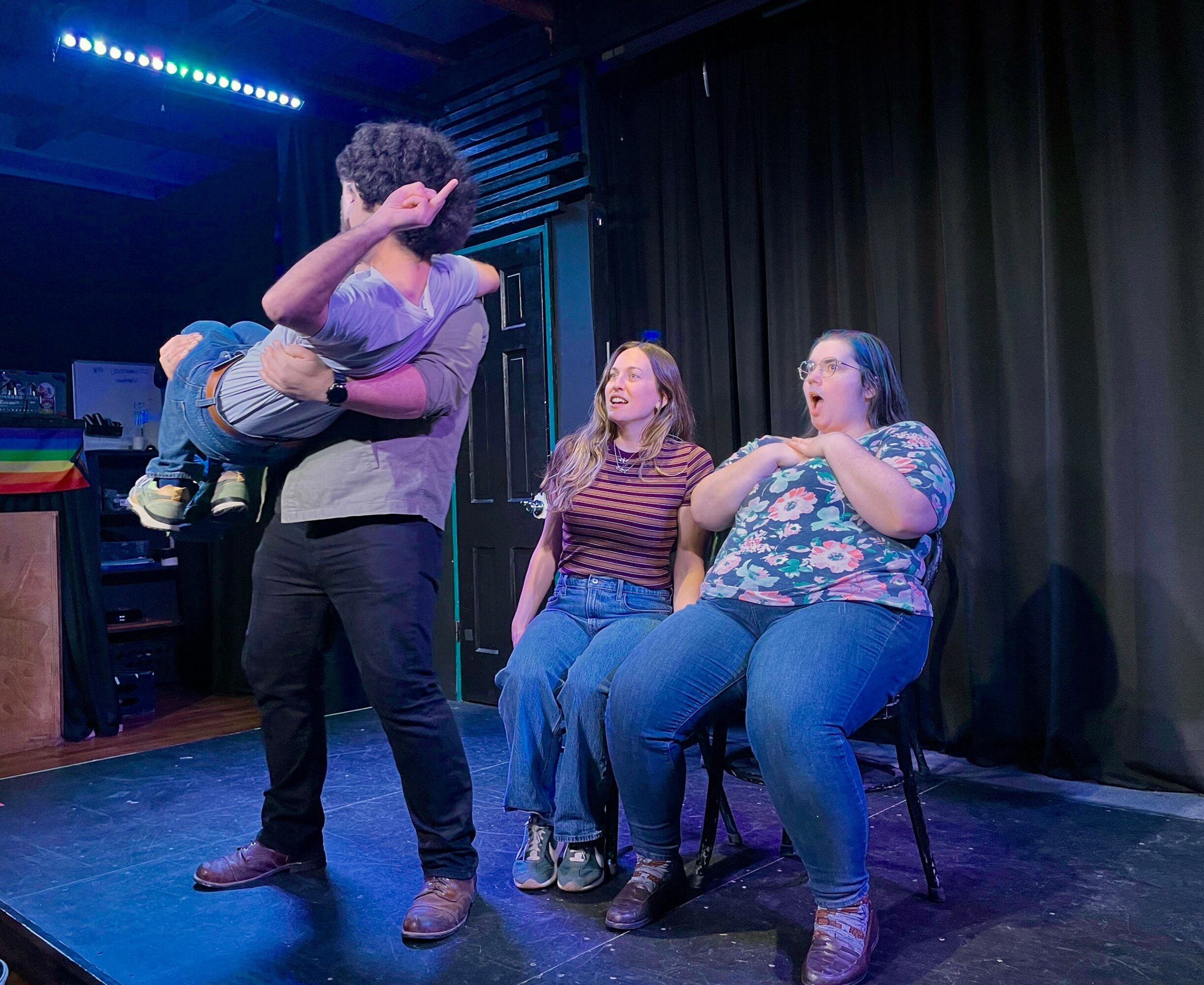 Person holding a child in a purple shirt, two women sitting and watching on stage, black curtain background.