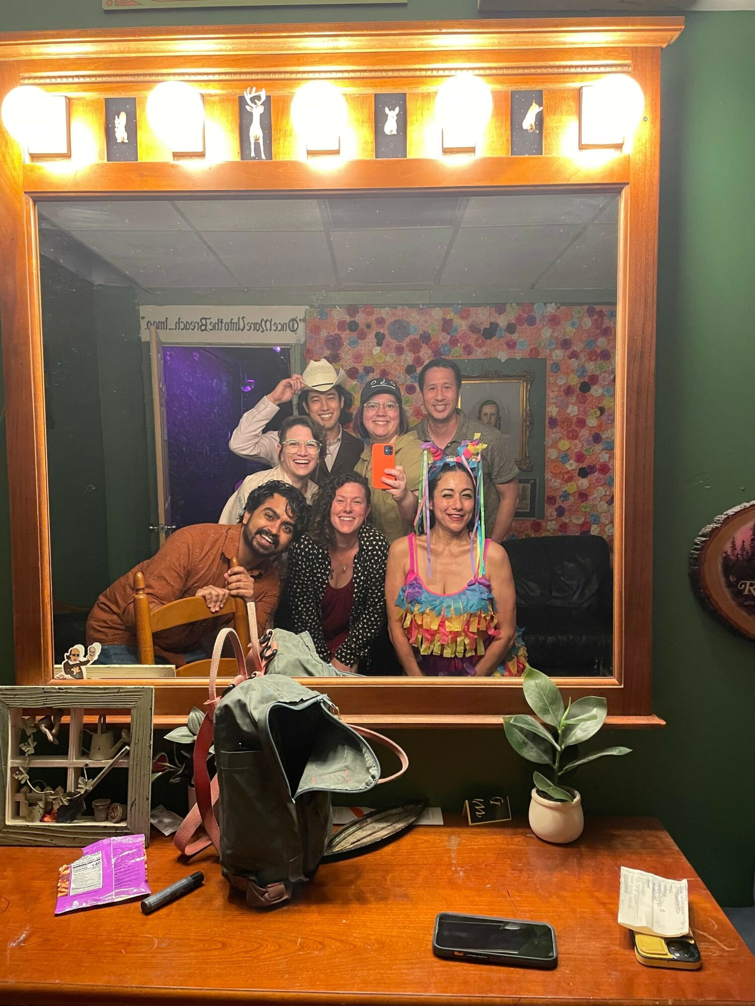 Group of five people in a room with floral wallpaper, some holding drinks, smiling, with a mirror and lights behind them.