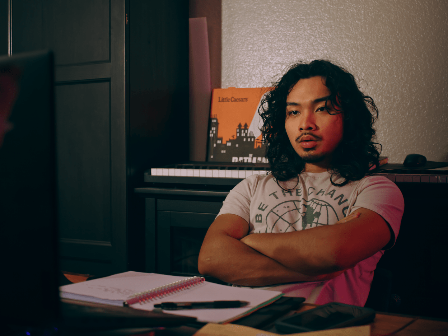 Person with curly hair sitting at a desk with a notebook and pen, in a room with a piano and books, arms crossed.