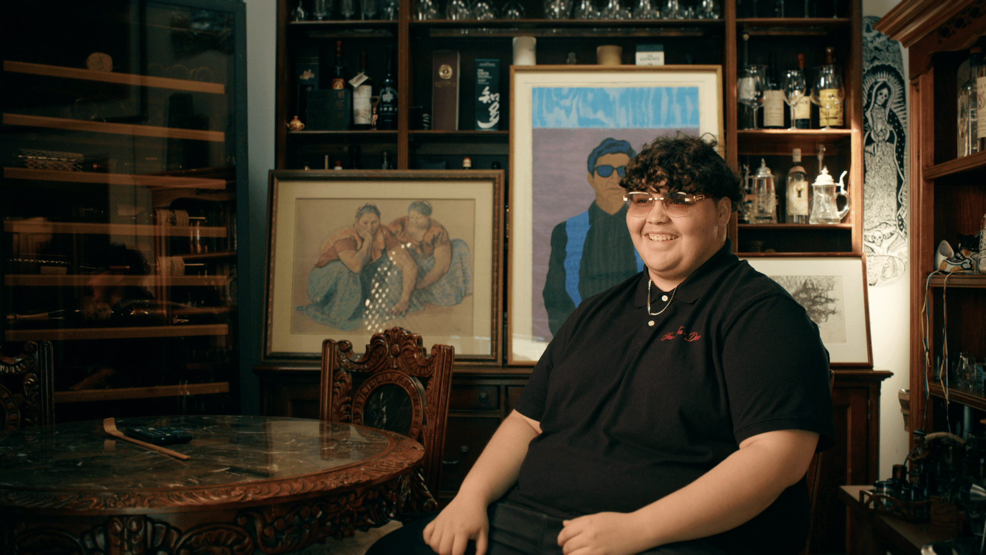 Person with curly hair and glasses sitting in a room with framed artwork and wooden furniture.