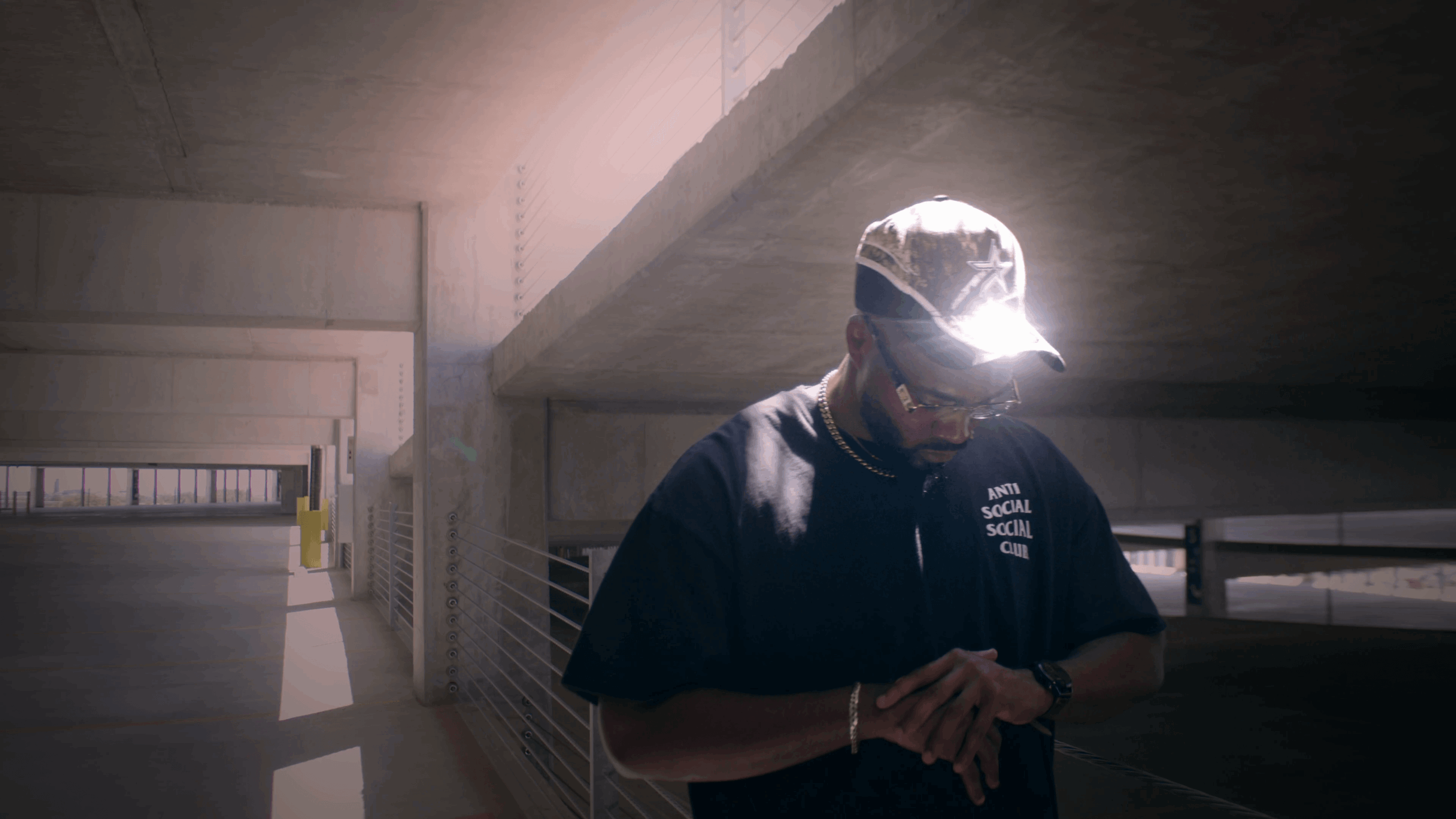 Person wearing a cap and dark shirt, standing under a concrete structure with sunlight behind them.