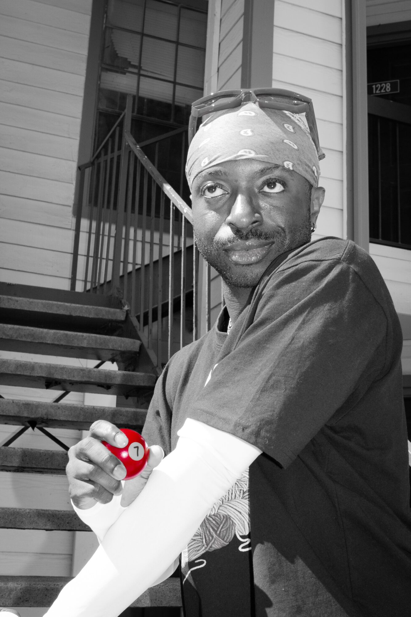Person wearing a bandana and glasses on head, holding a small object, standing outside a house with stairs and window.