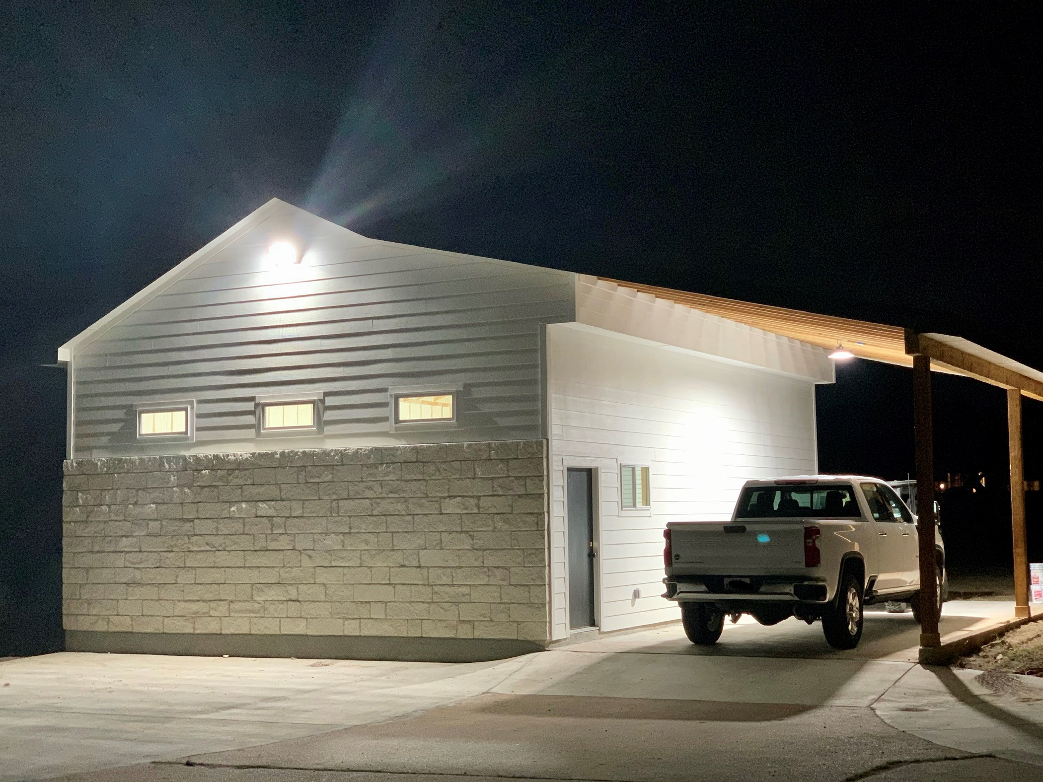 Nighttime view of a modern building with horizontal siding, a concrete block wall, and a parked vehicle under a wooden canopy.