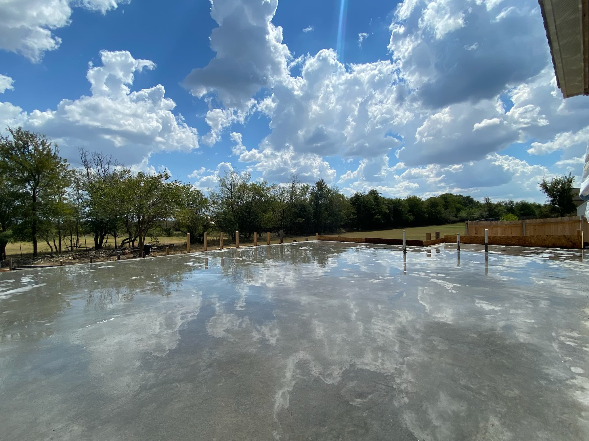 Sky with clouds reflected on a wet surface, trees and a fence in the background, part of a building roof visible.