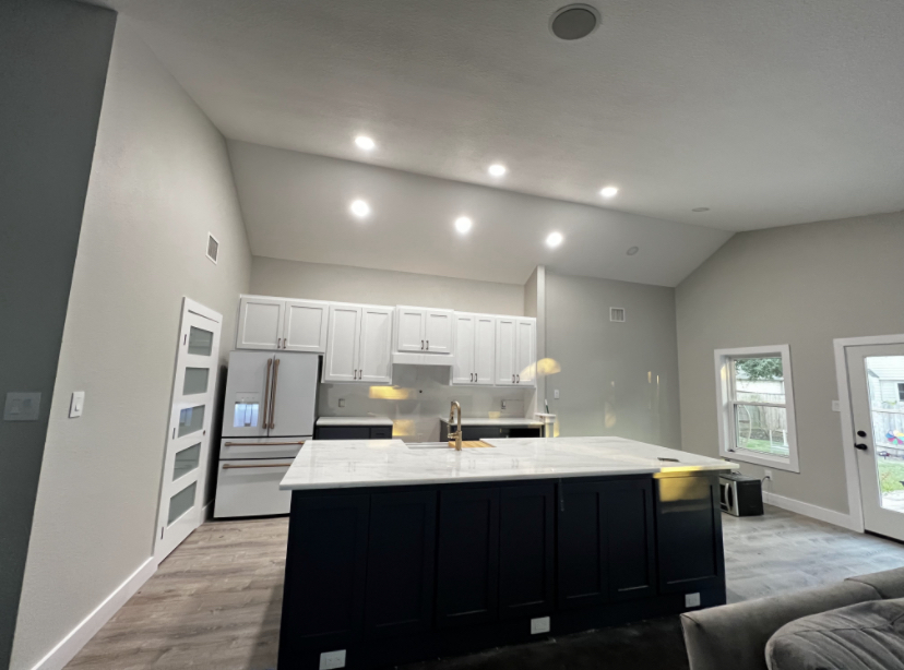 Modern kitchen with white cabinets, a large island with dark base, and stainless steel appliances, bright lighting, and windows.