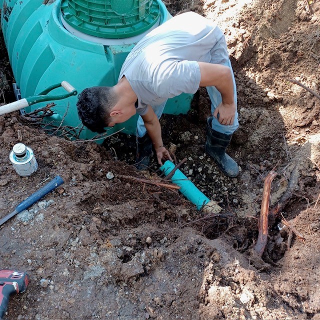 Person working in a trench with a large green tank nearby, using tools and wearing gloves and boots.