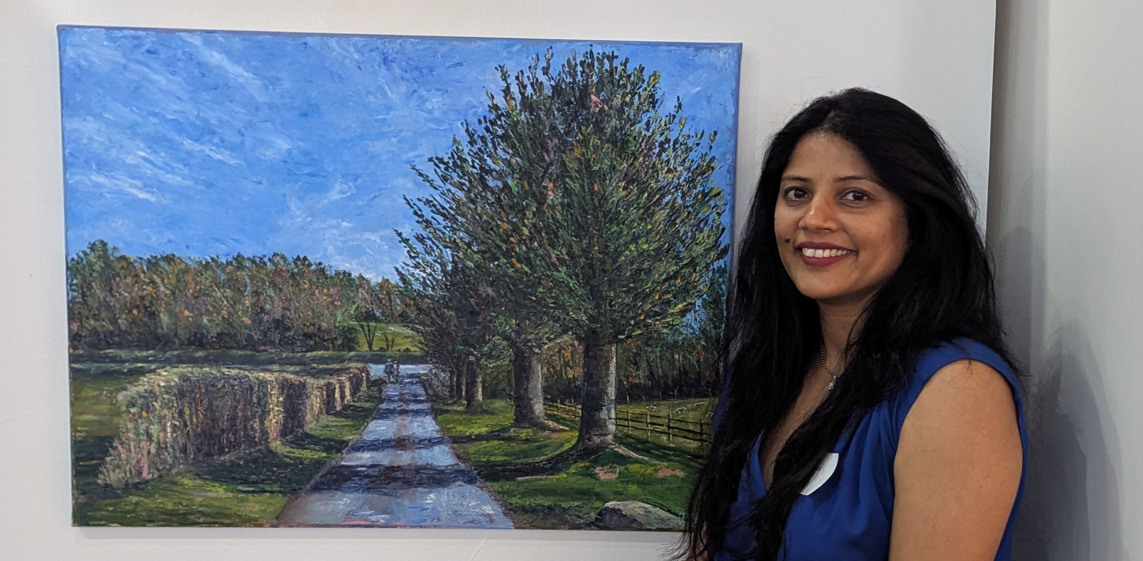 Woman with long dark hair smiling next to a landscape painting of a tree-lined path.