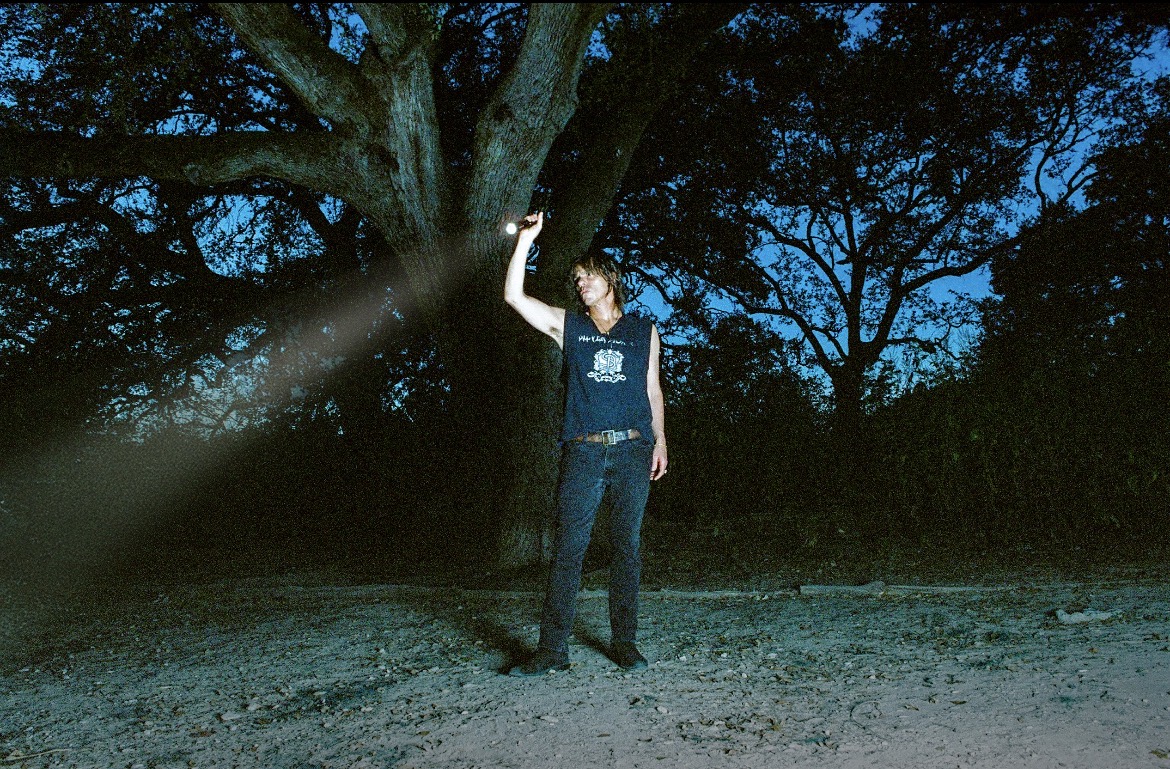 A person stands beside a large tree at dusk, holding a small object overhead as a beam of light crosses the scene.