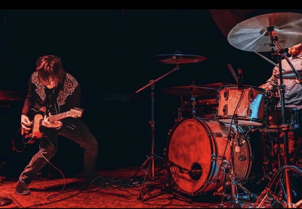 A guitarist plays beside a drum kit on a dark stage under red lighting.