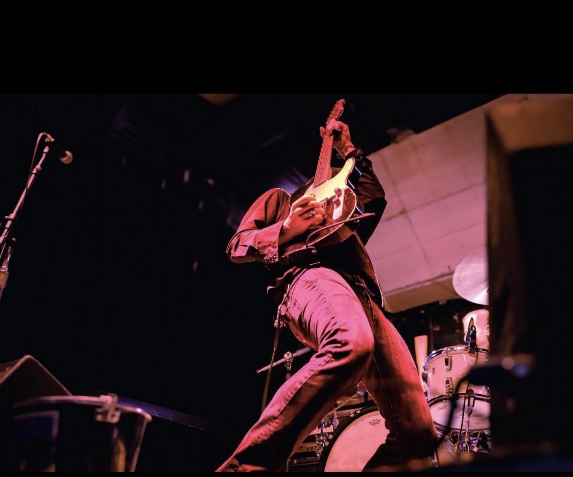 A musician plays an electric guitar onstage under red lighting, with a drum kit and microphone nearby.