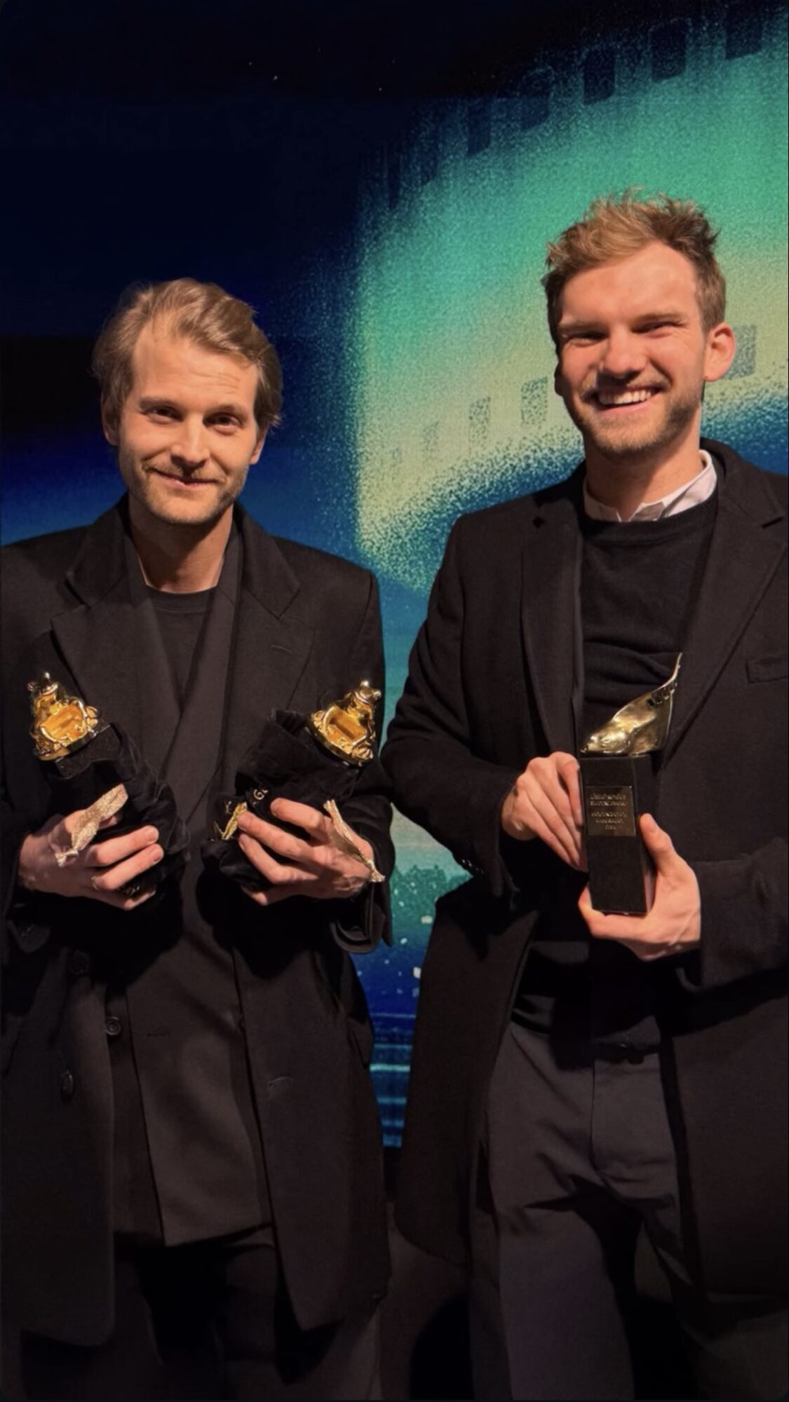 Two people in dark suits hold small trophy-like objects in front of a blue and green backdrop.