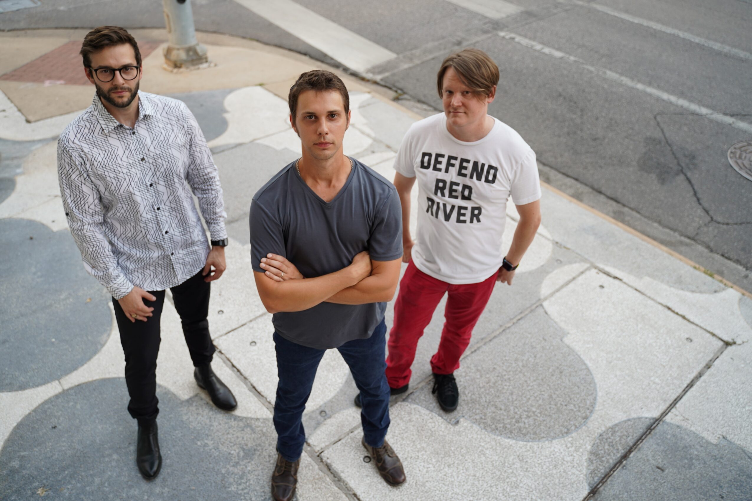 Three men standing on a sidewalk, looking up at the camera, with a street in the background.