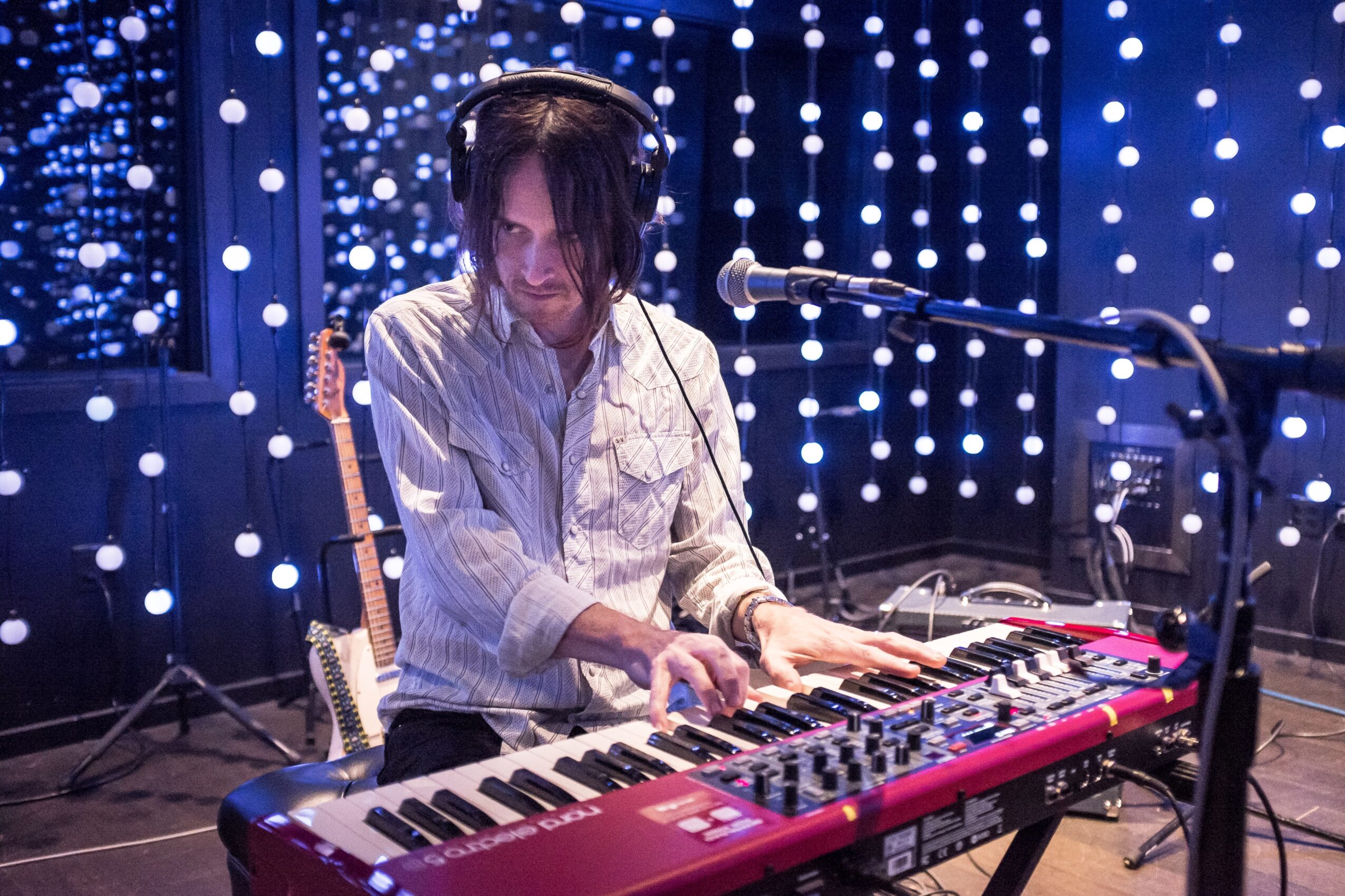 Person wearing headphones plays a red keyboard beside a microphone, with strings of lights in the background.