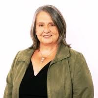 A person with shoulder-length hair wearing an olive jacket over a black top, standing against a white background.