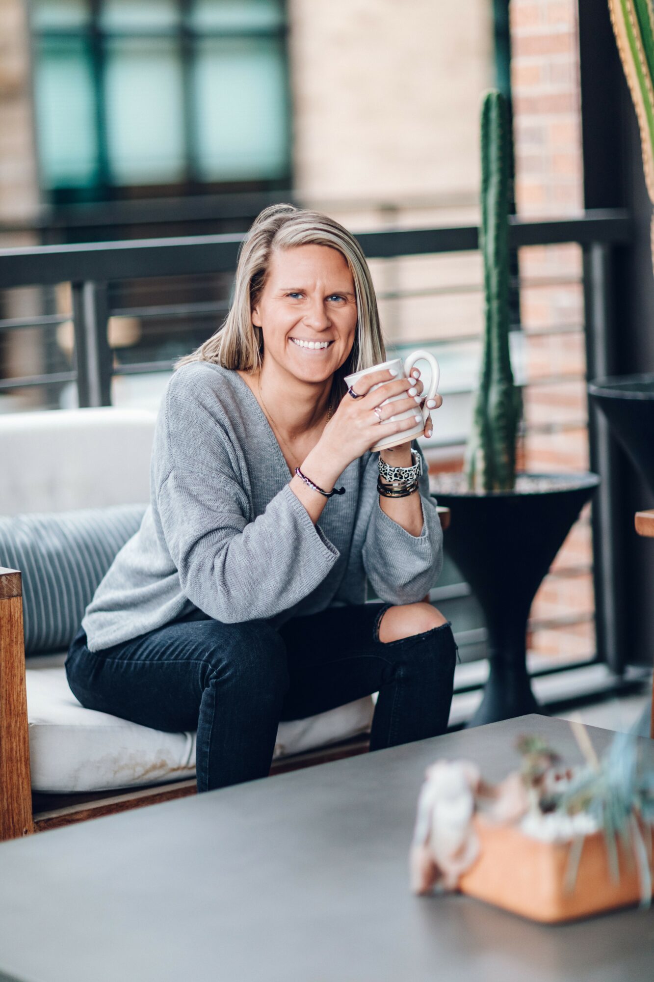 Woman sitting on a sofa holding a mug, smiling, in a bright room with large windows and plants.