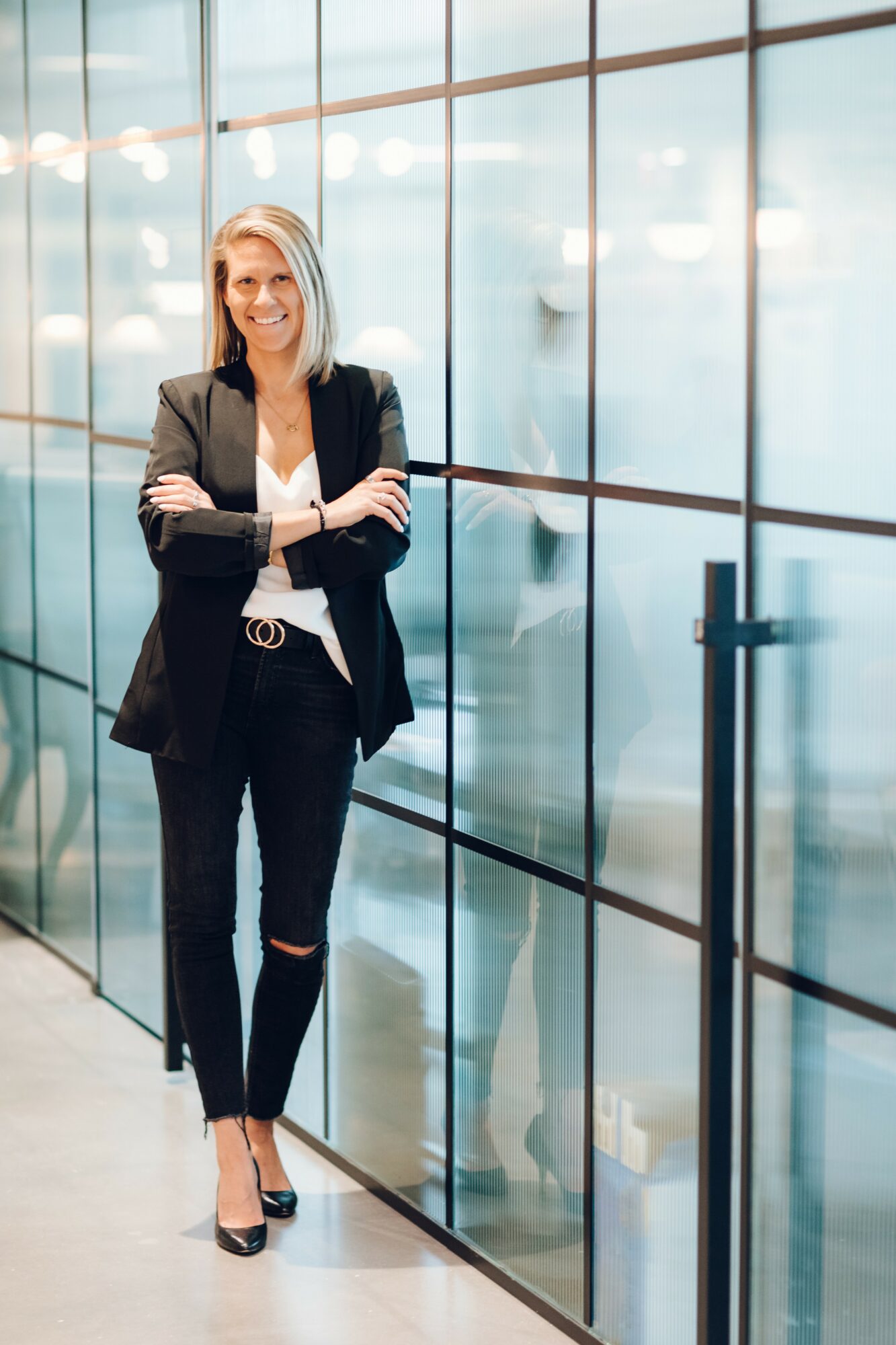 Woman with blonde hair in a black blazer and pants, standing with arms crossed in front of glass wall, smiling.