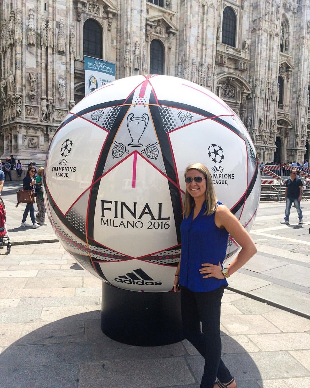 Woman standing next to a large soccer ball display in front of a historic building, wearing sunglasses and a blue top.