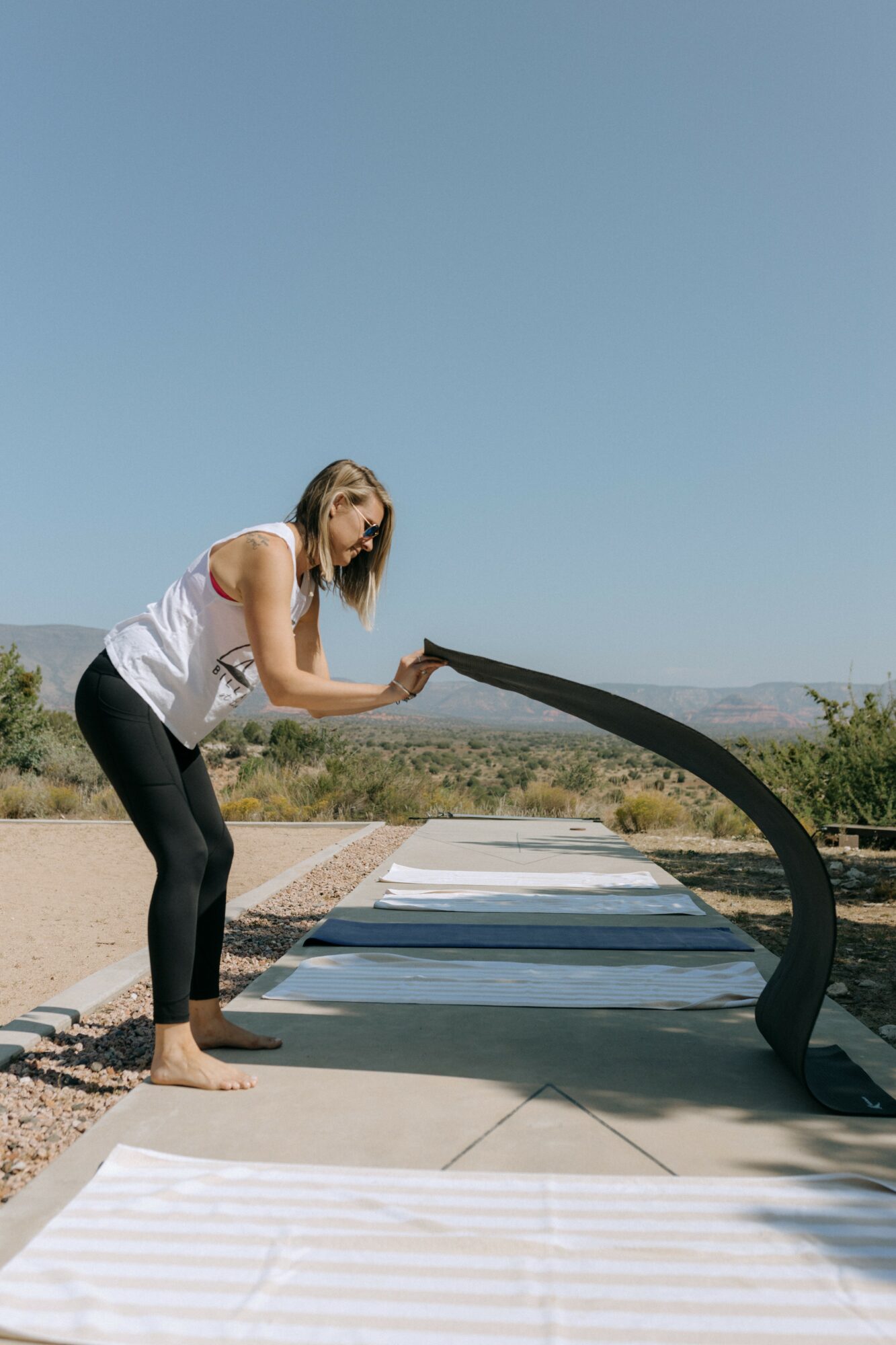 Woman in white top and black pants bending over outdoor lounge chairs under clear blue sky.