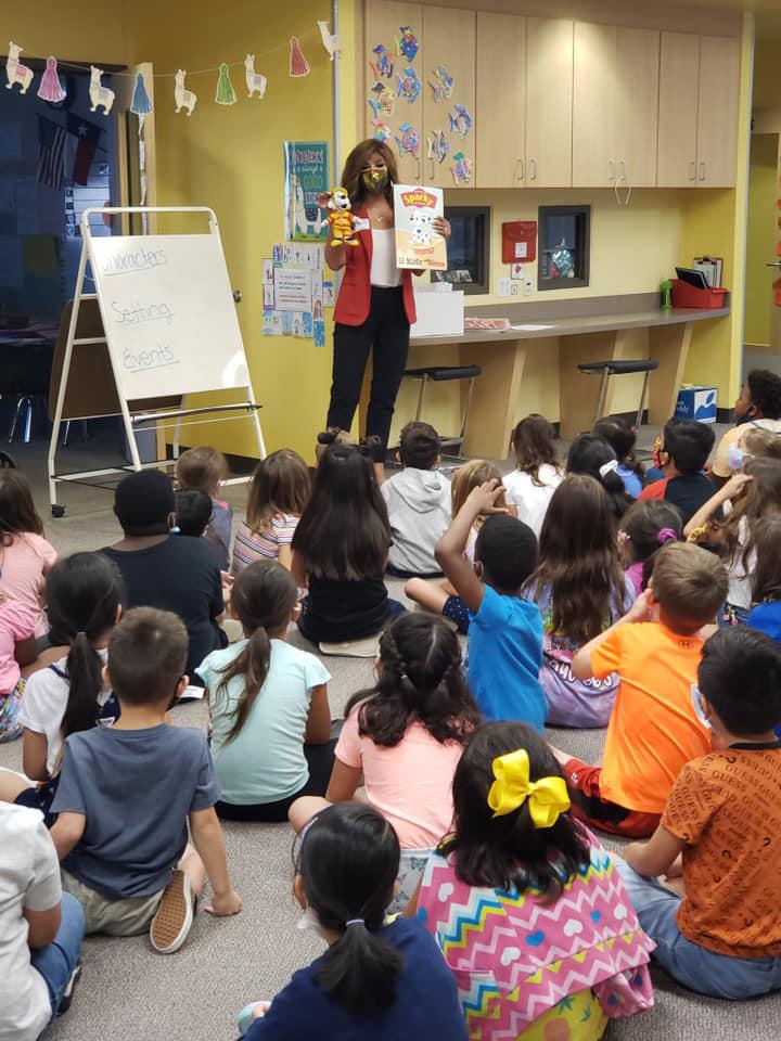 Teacher standing in front of children in a classroom, holding a book and engaging students during a lesson.