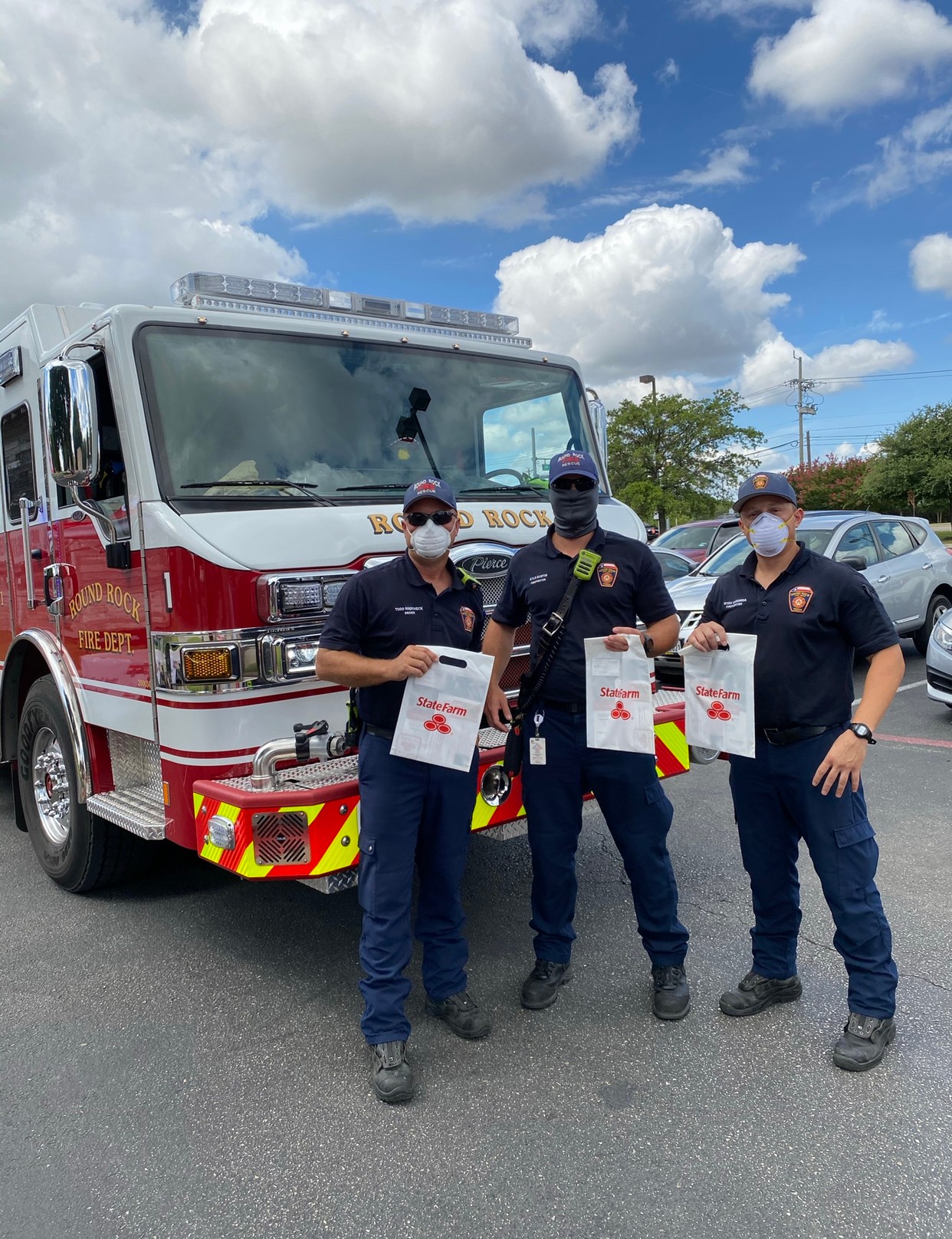 Three firefighters in uniform and masks stand in front of a fire truck holding papers, outdoors on a partly cloudy day.