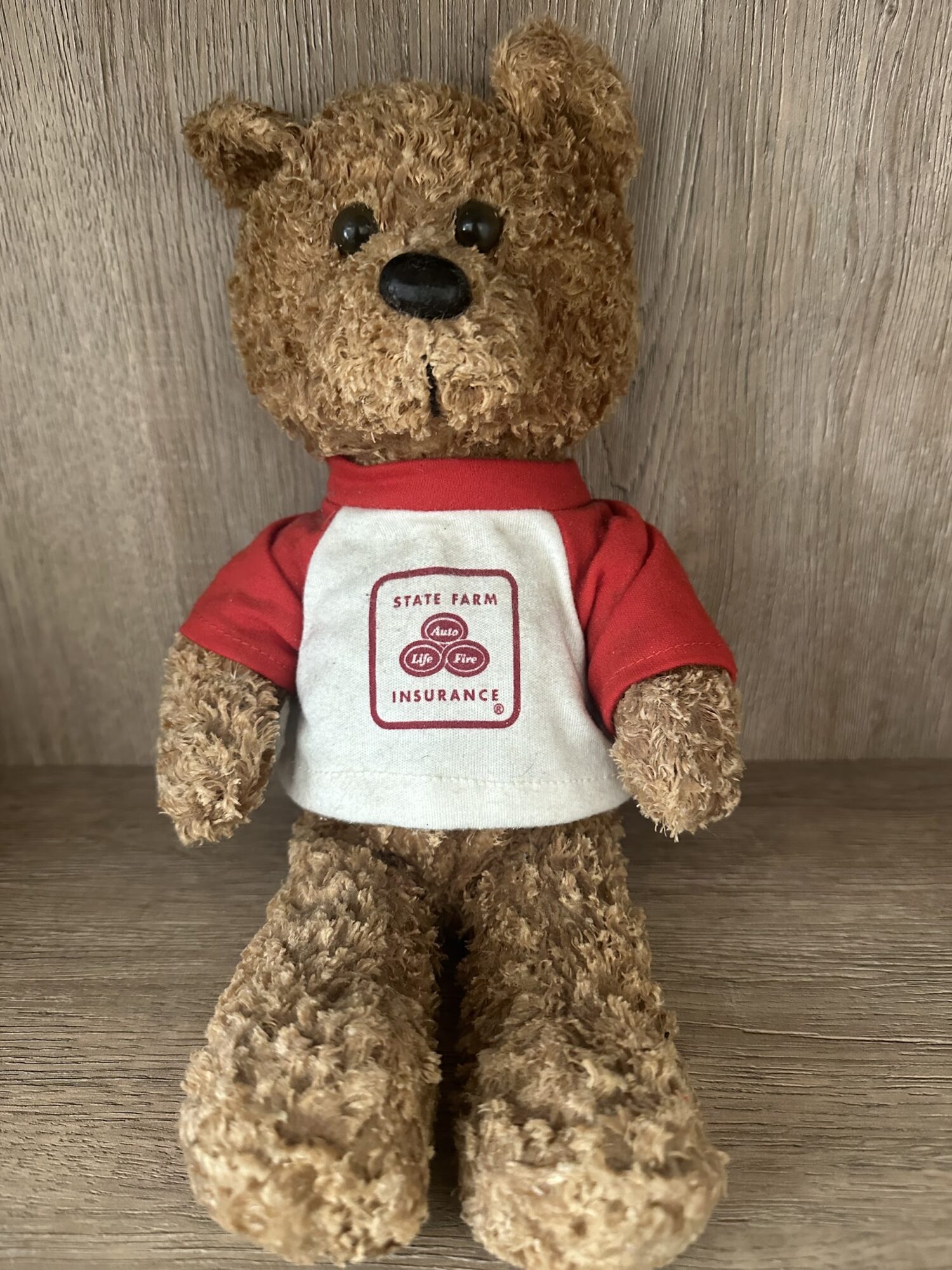 Stuffed bear wearing a white and red shirt with a logo, standing on a wooden surface against a wooden background.