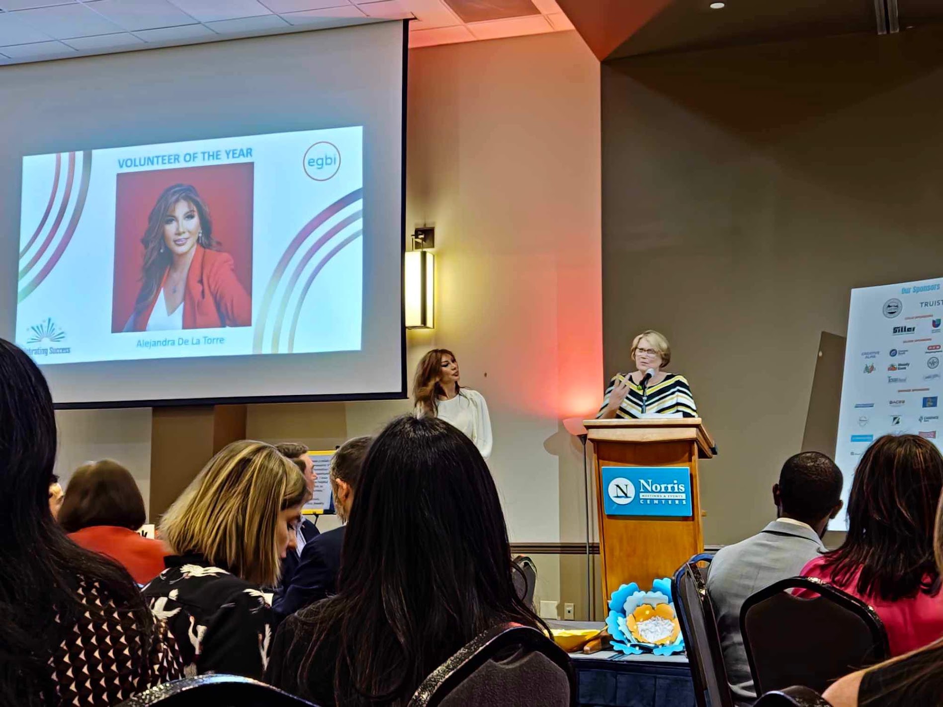 Two women speaking at a podium in front of an audience, with a large screen displaying a woman in a red outfit and text.