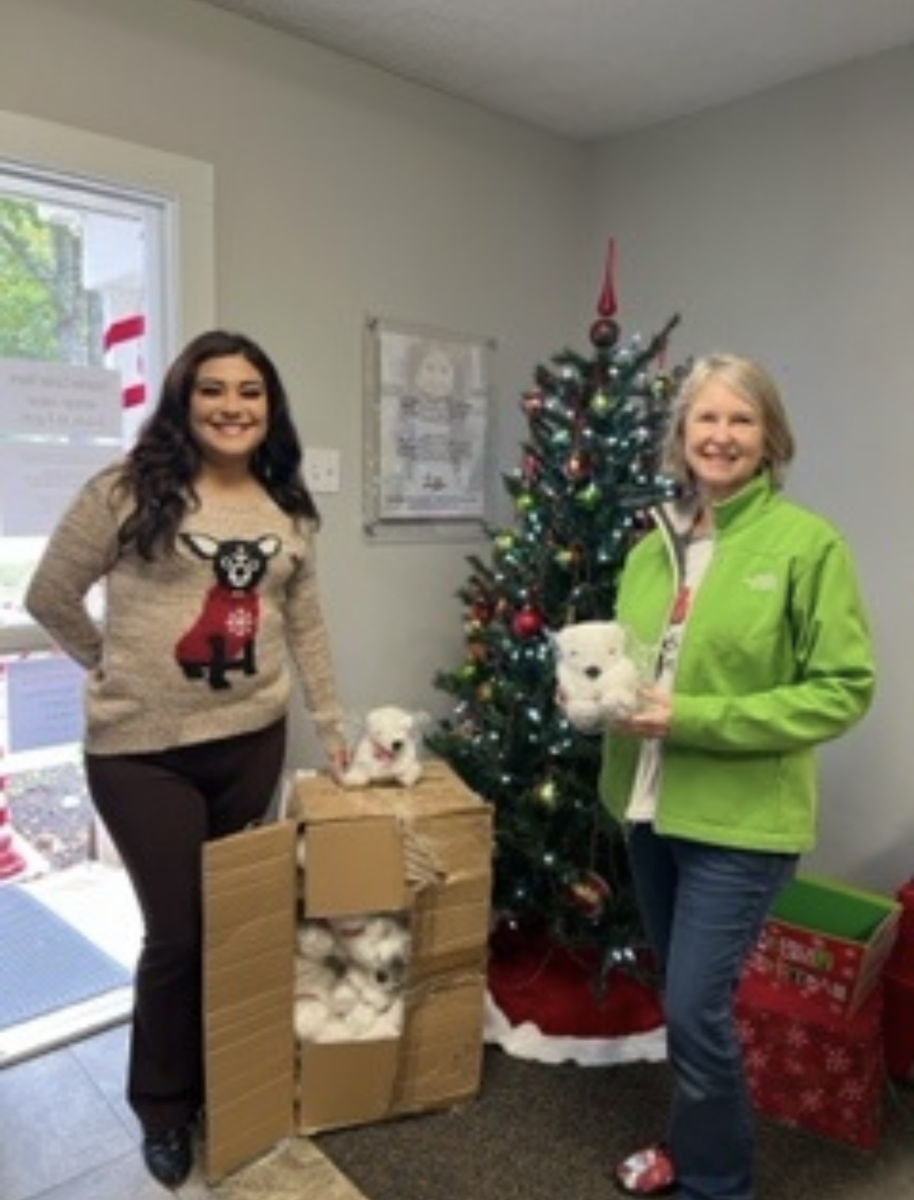 Two women stand next to a decorated Christmas tree, holding a stuffed animal, with boxes of ornaments nearby.
