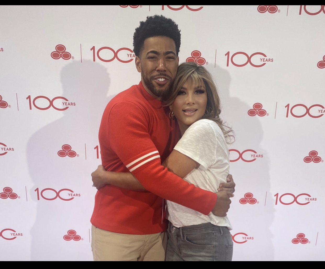 Two people hugging in front of a backdrop with red and white logos and text, smiling at the camera.