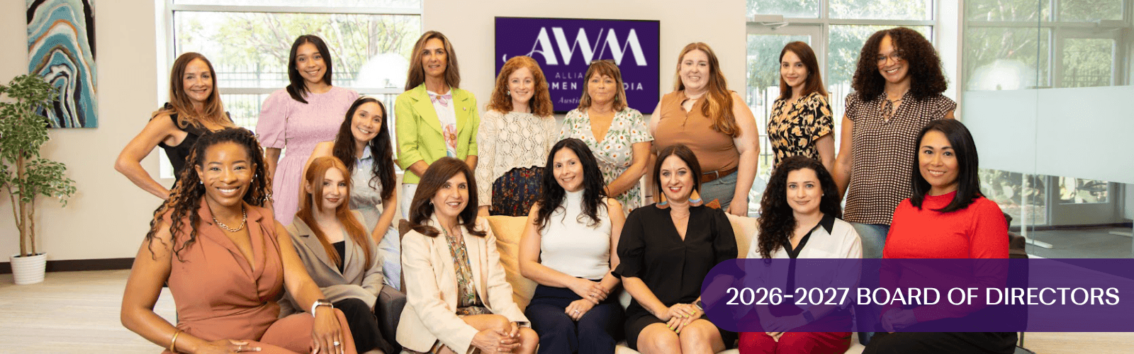 Group of women sitting and standing in a room with a purple banner, smiling, with large windows in the background.