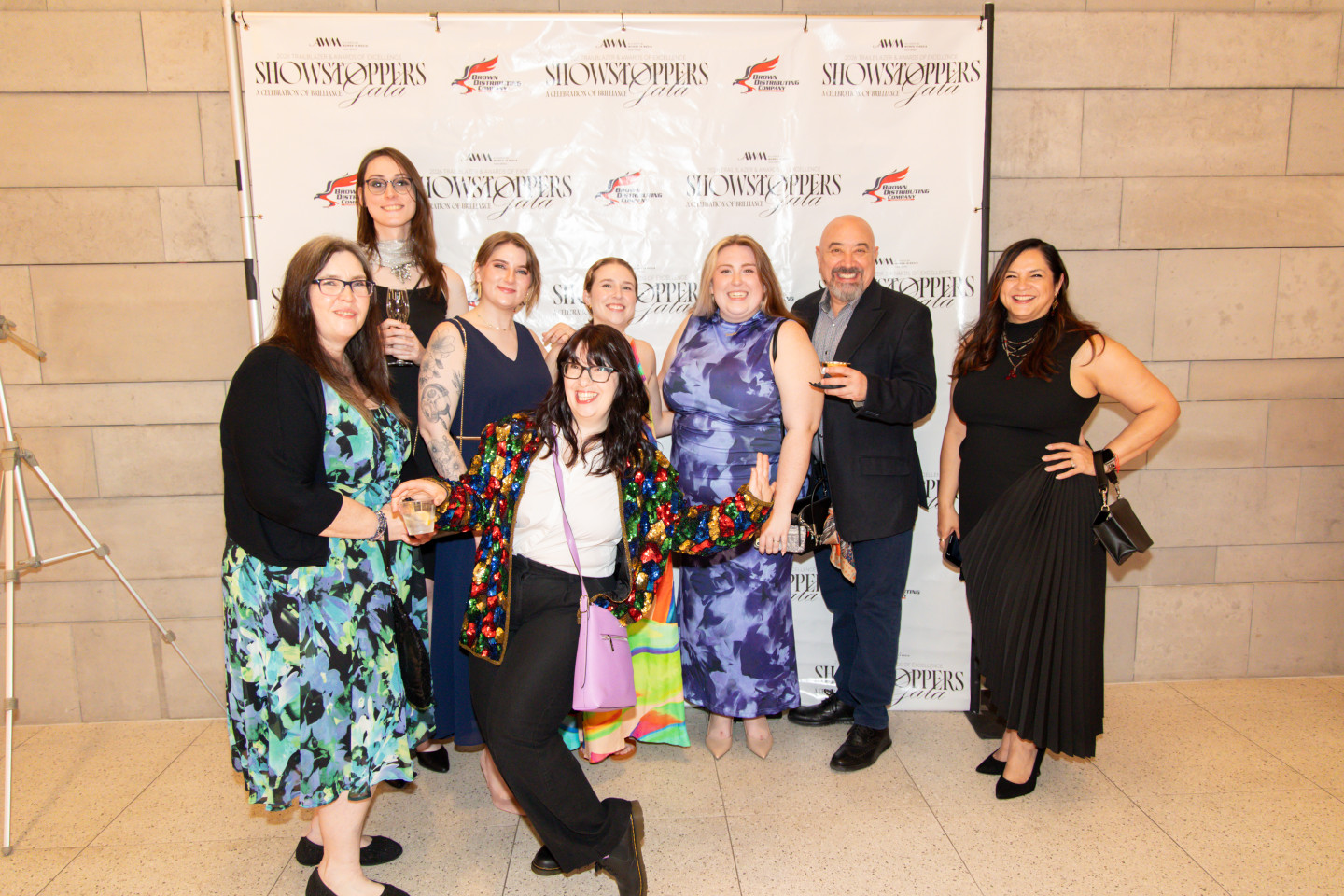 Seven people pose in front of a backdrop with logos, standing on a tiled floor against a stone wall.