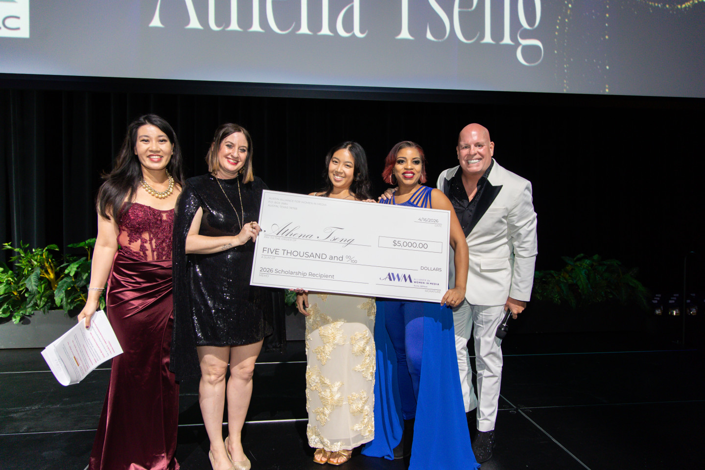 Group of five people on stage holding a large check, smiling, with a dark background and a screen above them.