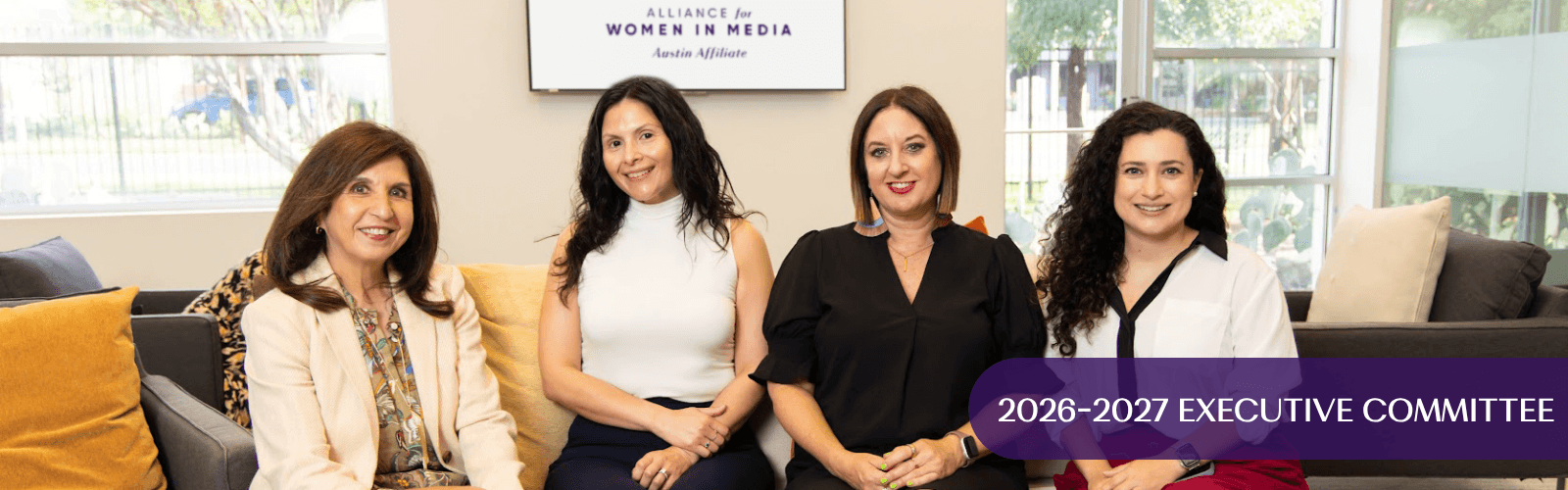 Four women sitting on a sofa in a room with large windows, smiling, with a sign reading 2026-2027 Executive Committee above them.