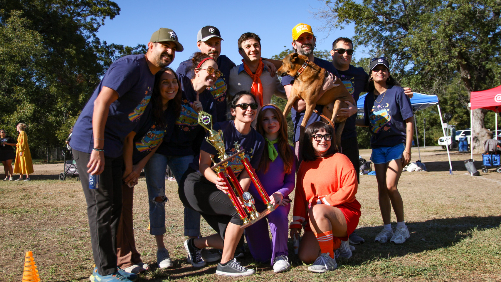 Group of people outdoors, some holding a trophy, smiling, with trees and tents in the background.