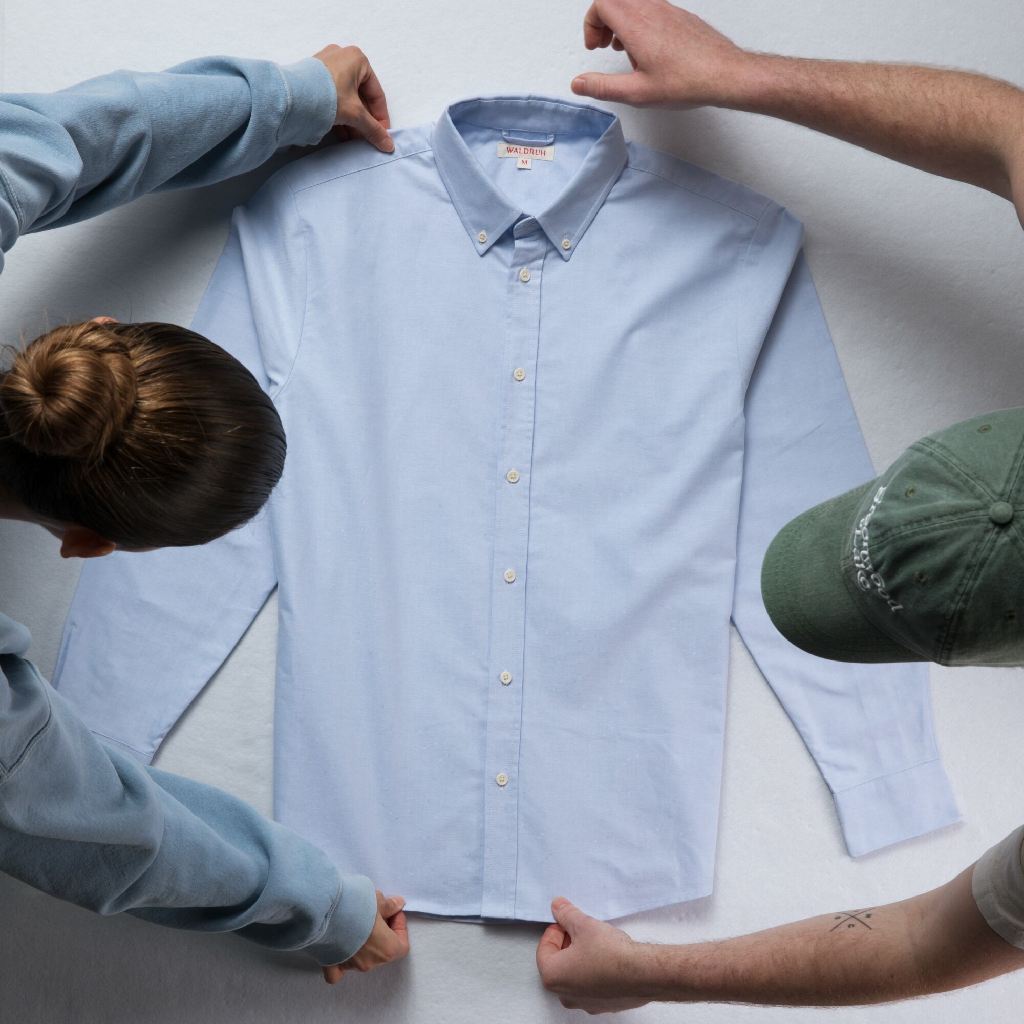 Three people hold a light blue button-up shirt on a white surface, viewed from above, with one person wearing a green cap.